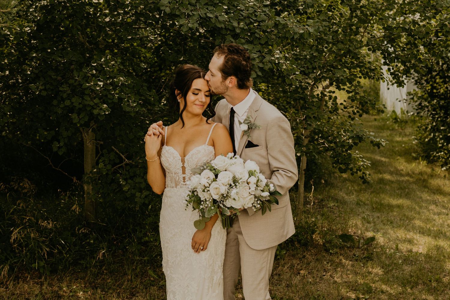 Newlyweds share intimate moment with white floral bouquet against green foliage.