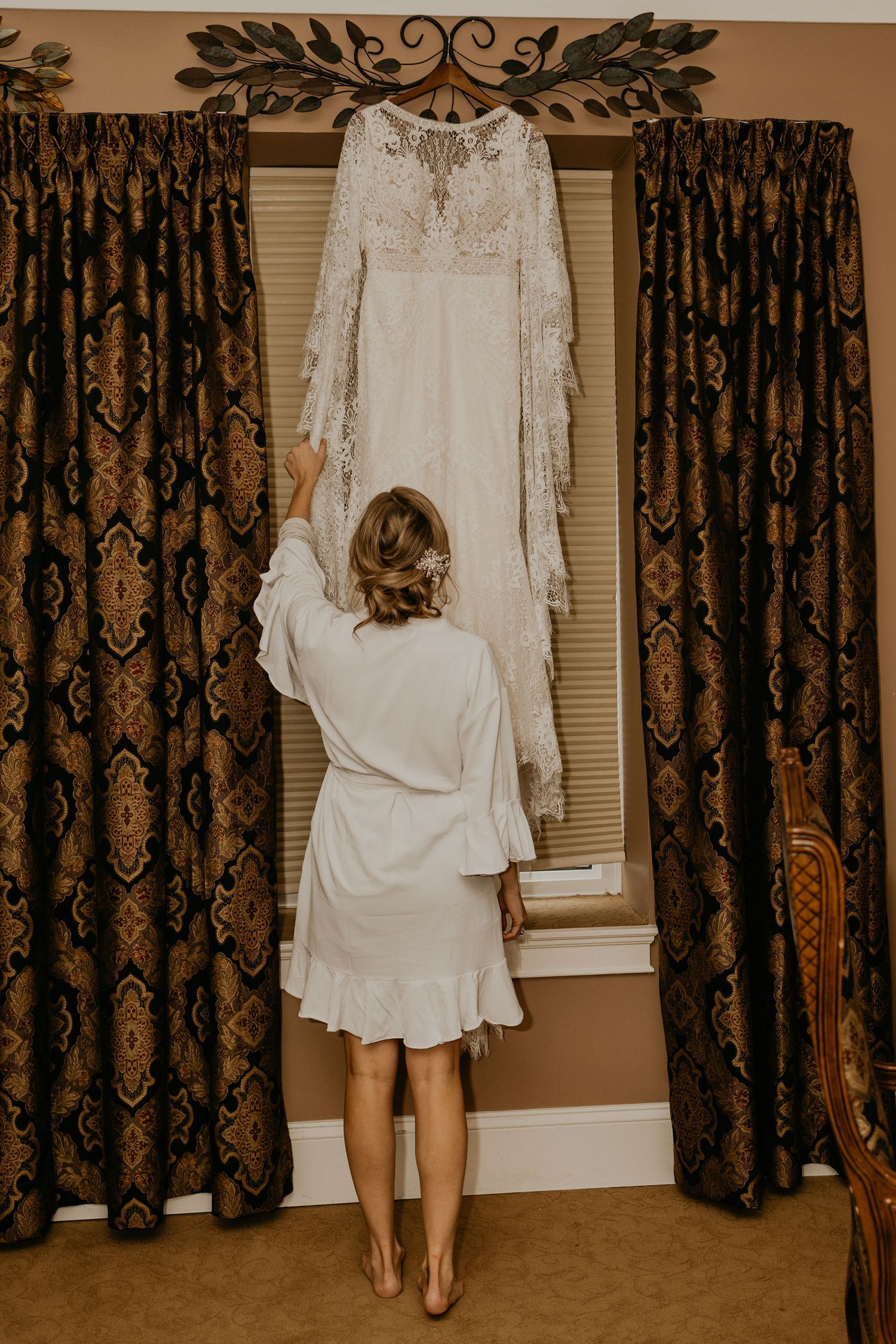 A woman in white robe admires her wedding dress hanging by a window.