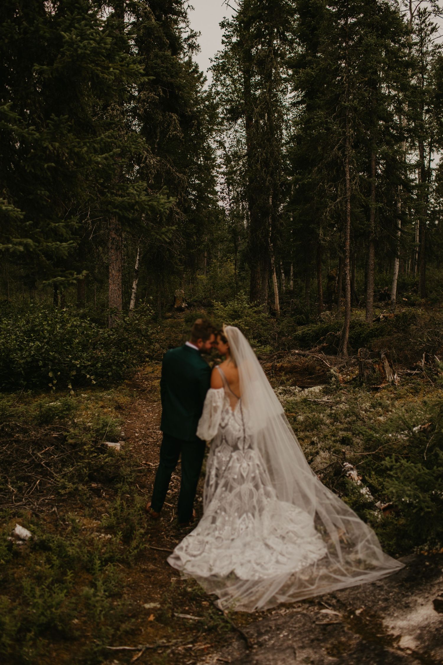 Dramatic forest wedding portrait with flowing lace dress and emerald suit.