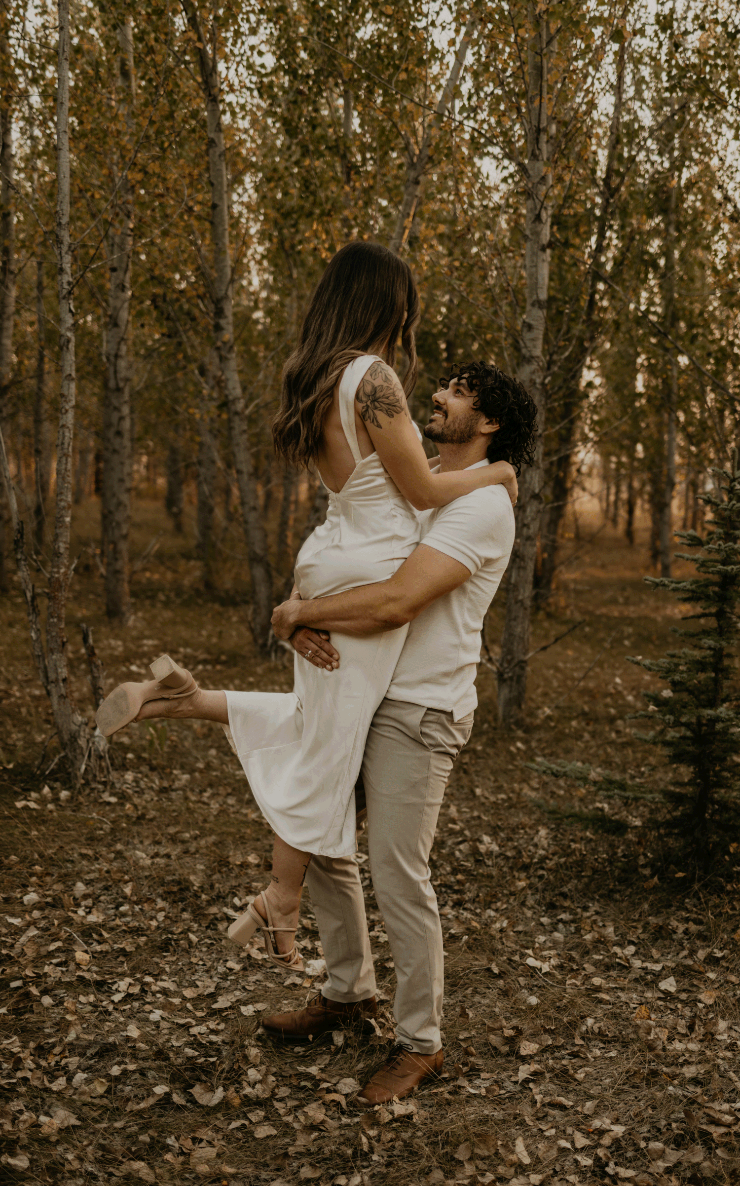 A romantic couple embraces in a golden autumn forest, with the person in white lifting one foot off the ground.