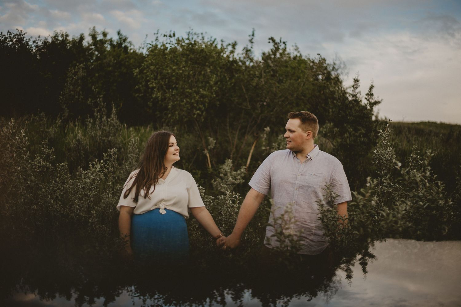 A couple holds hands while standing in a field of tall grass and wildflowers at sunset.