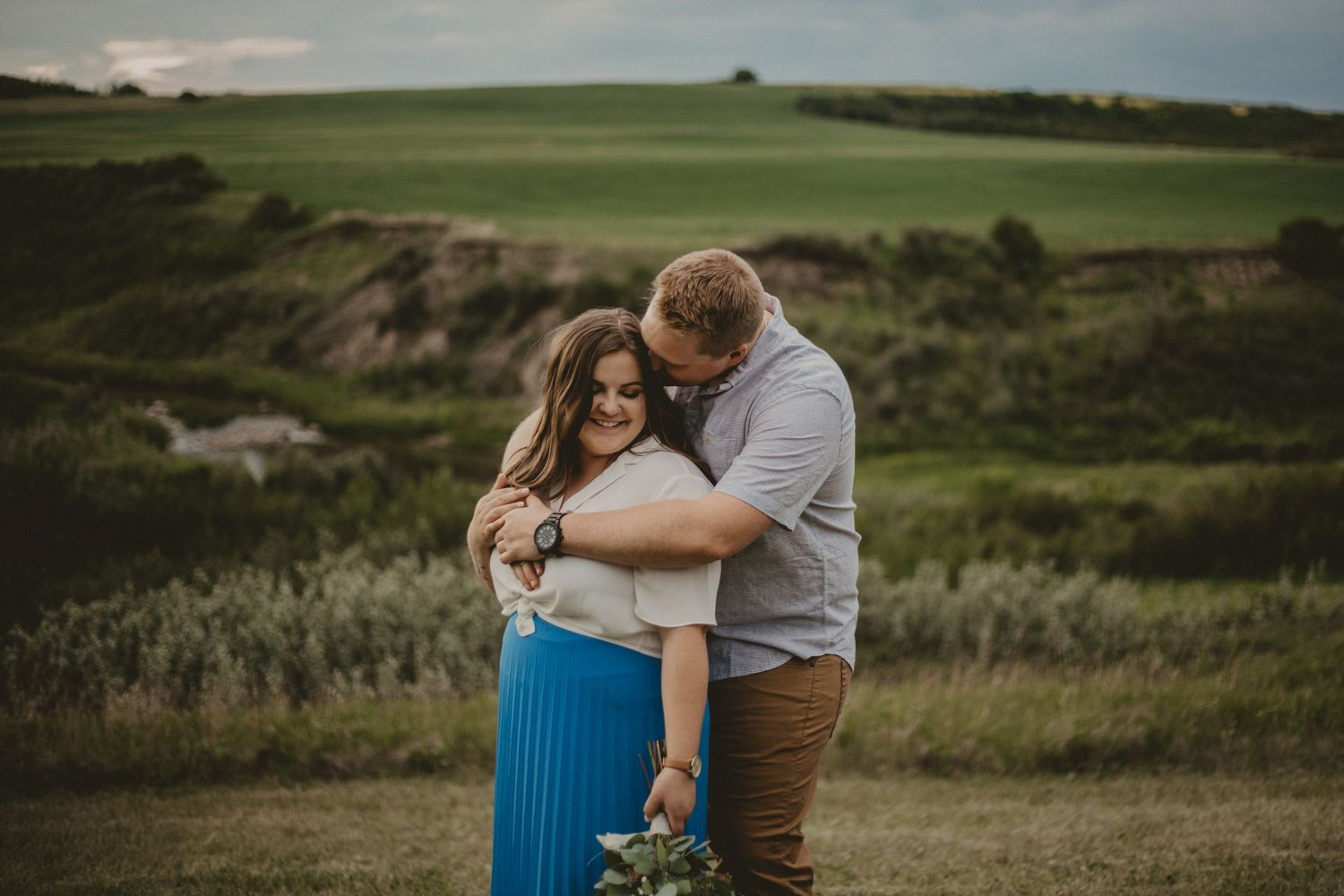 Intimate outdoor portrait of couple embracing on grassy hillside at sunset.