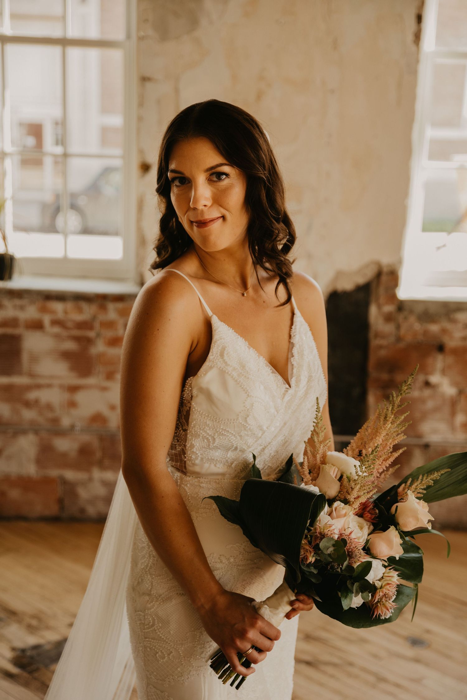 A bride in a fitted lace wedding gown holds a beautiful bouquet in a rustic venue.