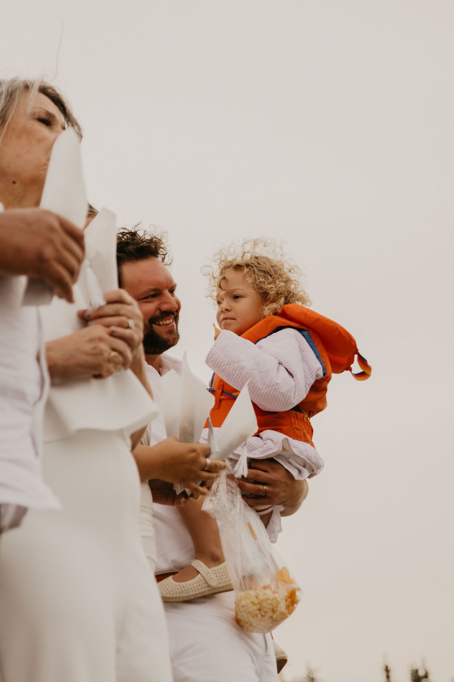 A family celebration outdoors with a small child wearing an orange jacket being held against a bright background.