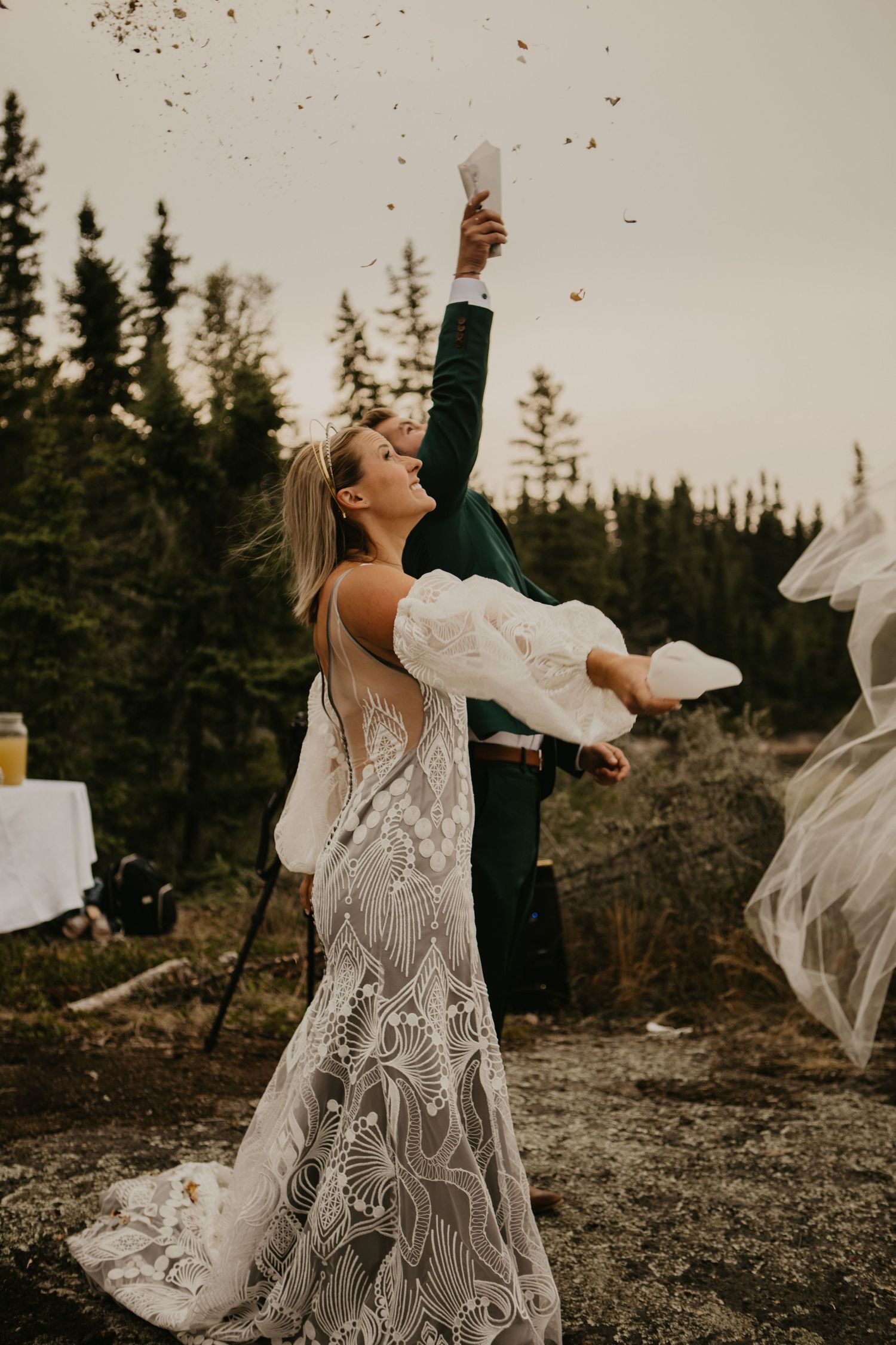 A bride in a detailed lace gown celebrates joyfully outdoors with pine trees in the background.
