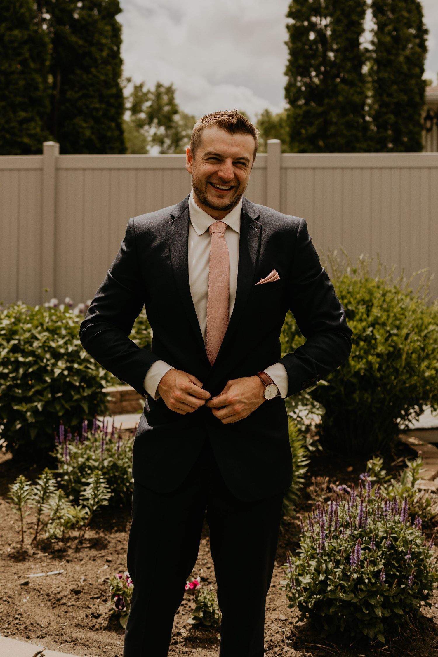 Wedding fashion portrait showing groom in dark suit with pink tie in garden setting.