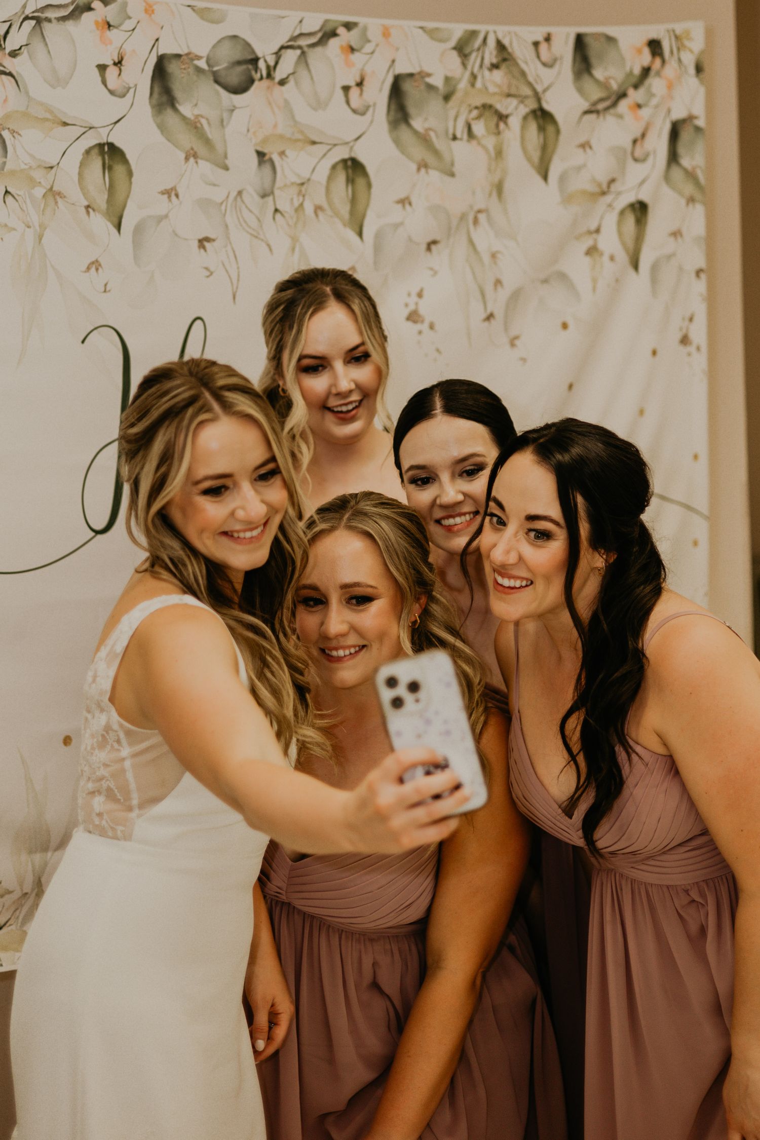 Friends taking selfie at wedding against floral backdrop.