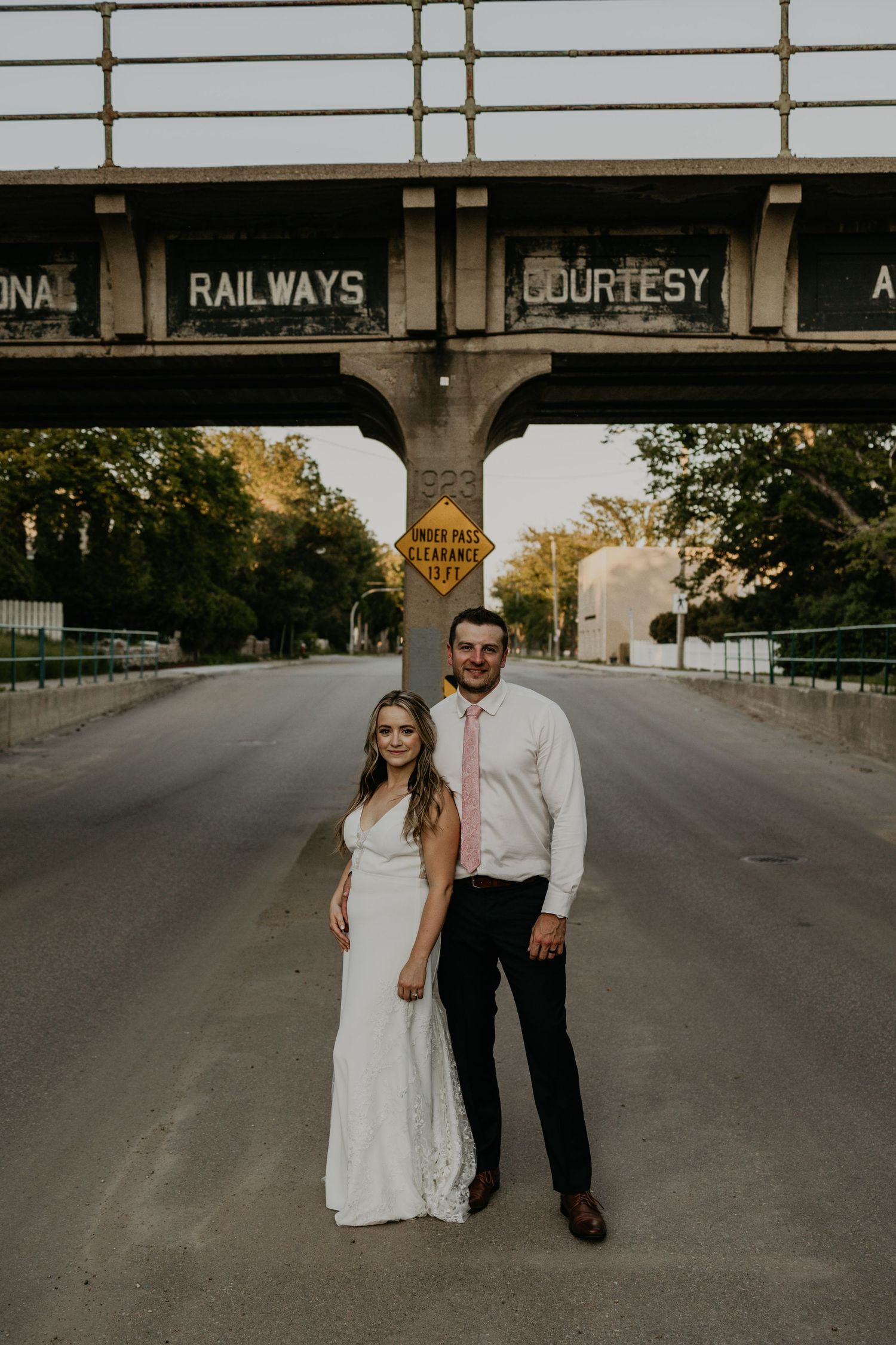 A couple poses for a photo under a railway bridge with 'COURTESY' signage during a wedding photoshoot.