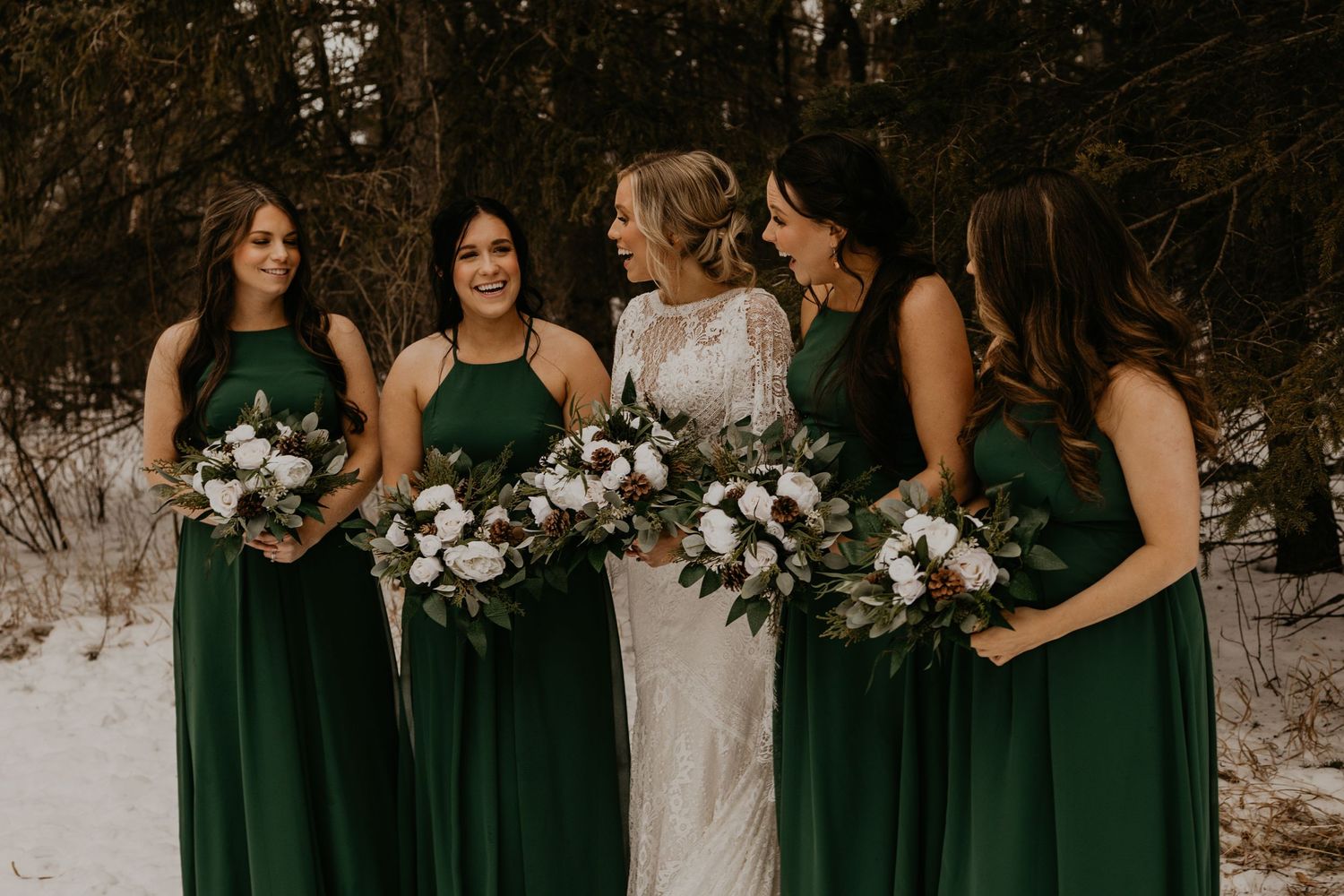 Group portrait of bridal party in emerald dresses holding white floral bouquets against winter forest.