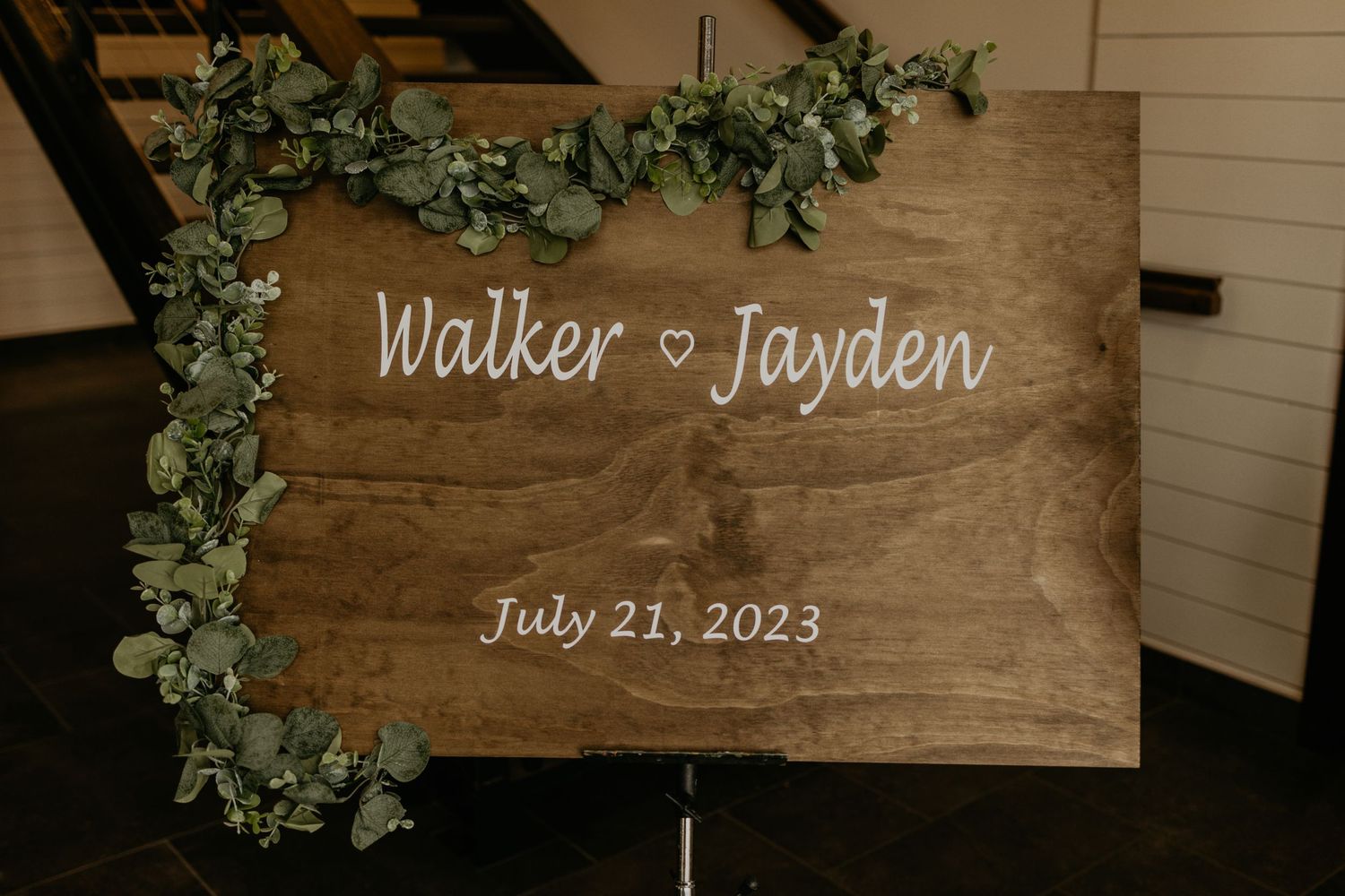 Rustic wooden wedding sign with greenery decoration displaying names and date.