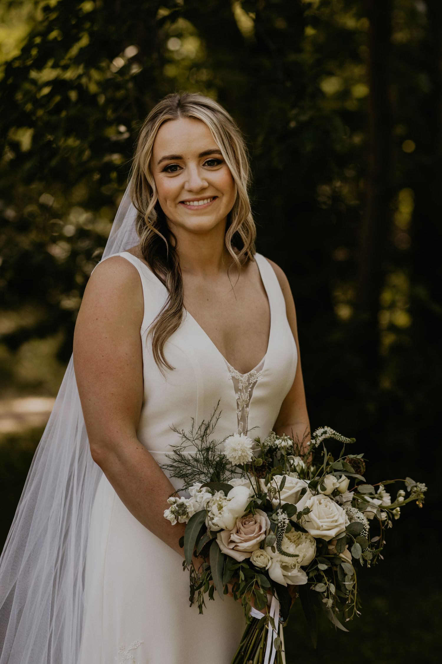 Bride holds elegant white and blush bouquet while smiling in natural outdoor setting.
