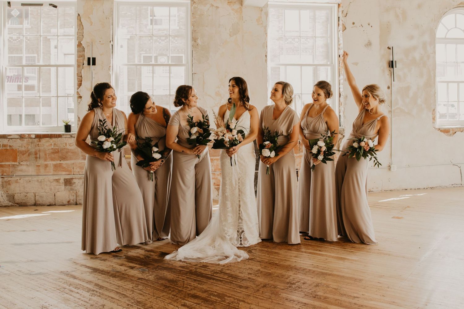 Bridal party in matching beige dresses poses with flowers in front of bright windows.