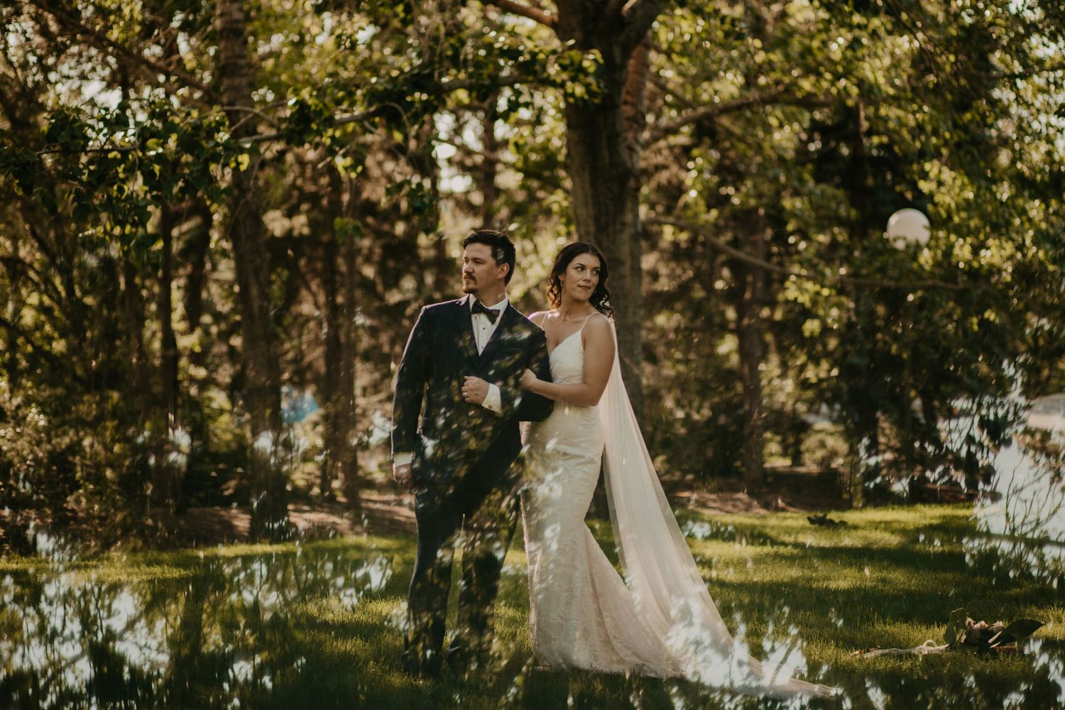 Wedding couple walks through sun-dappled garden with elegant white dress and long veil.