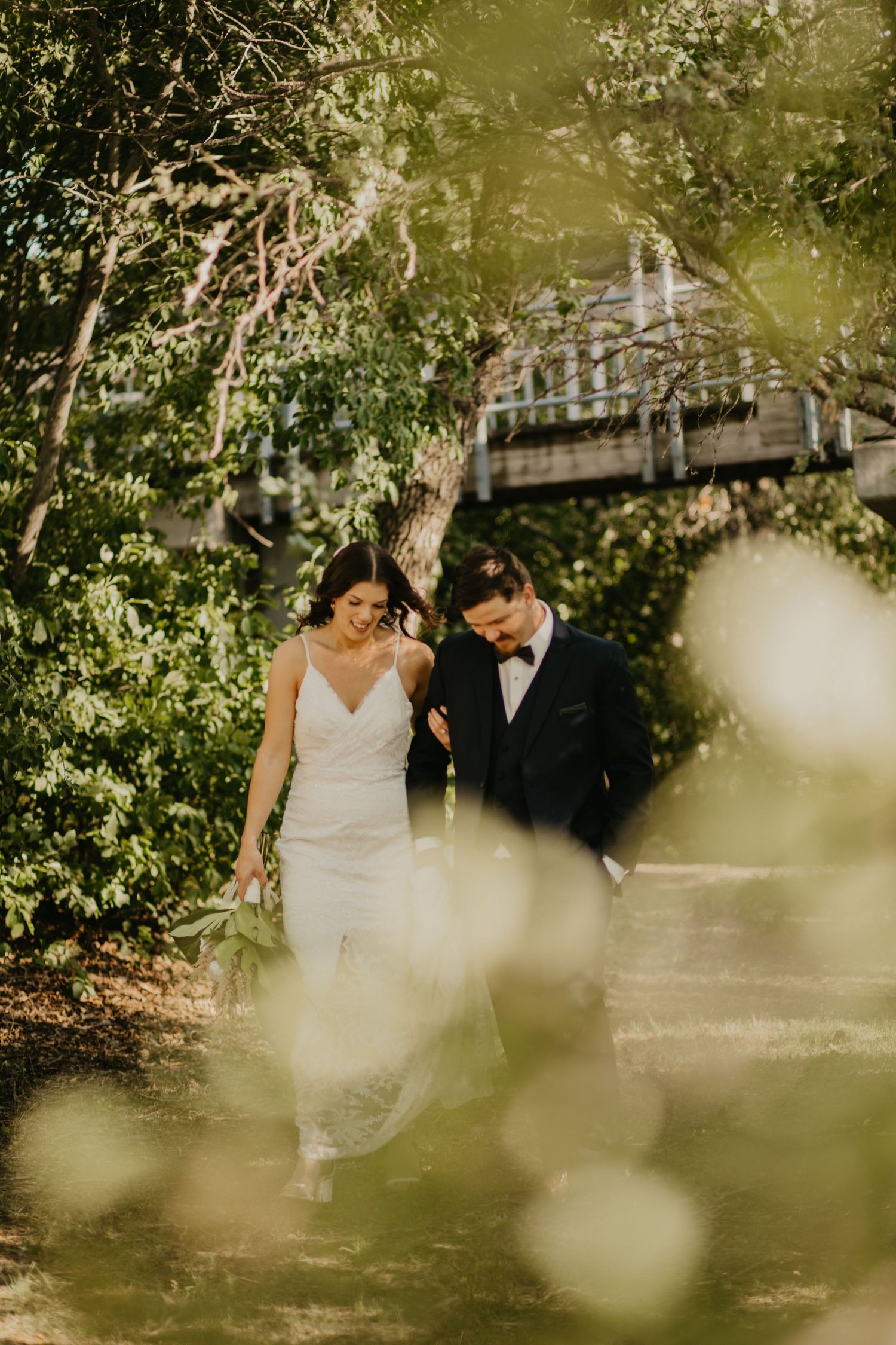 Couple walking through garden path with wooden bridge visible through bokeh effects.