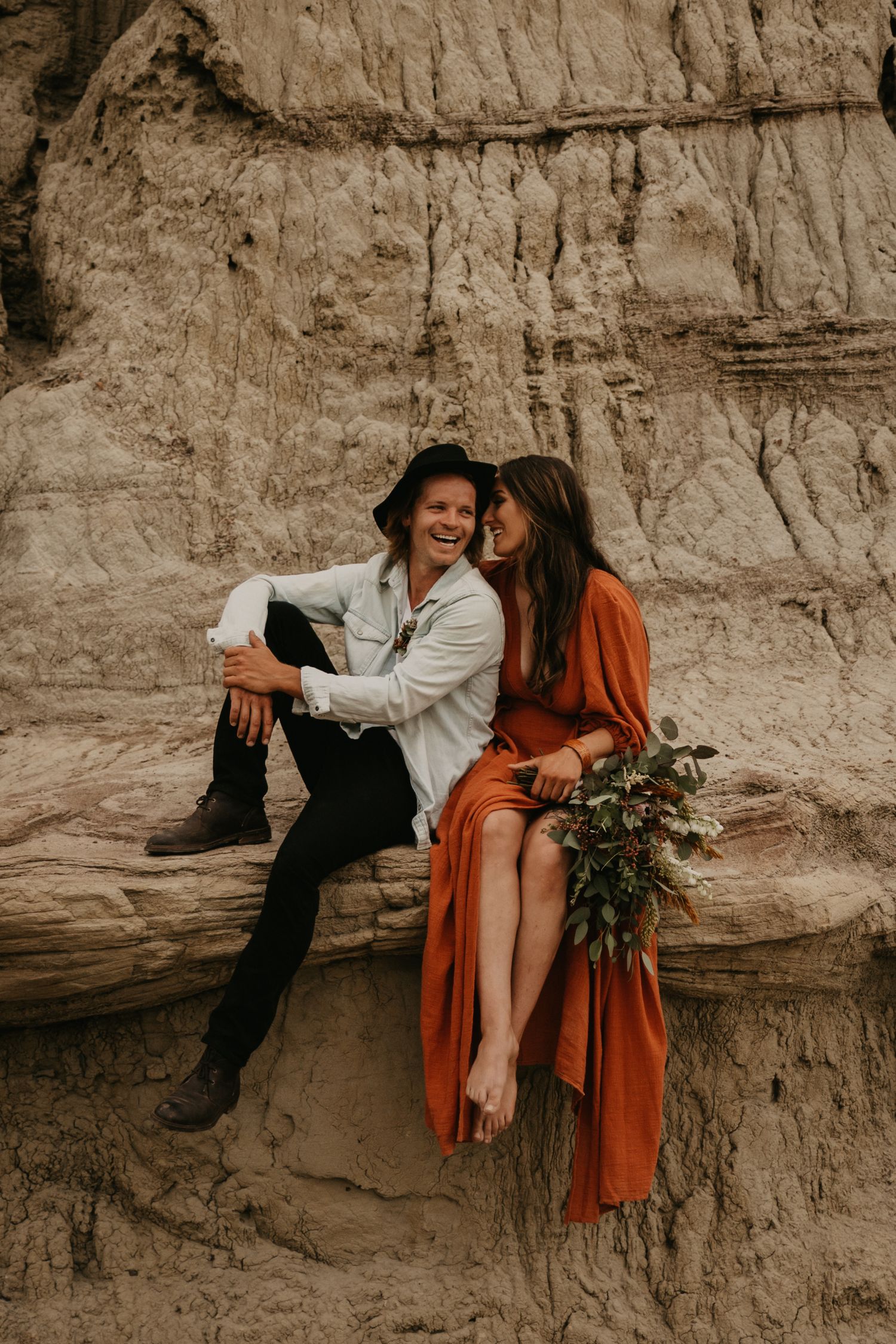 A couple relaxing on a rocky ledge against weathered cliff walls in bohemian attire.