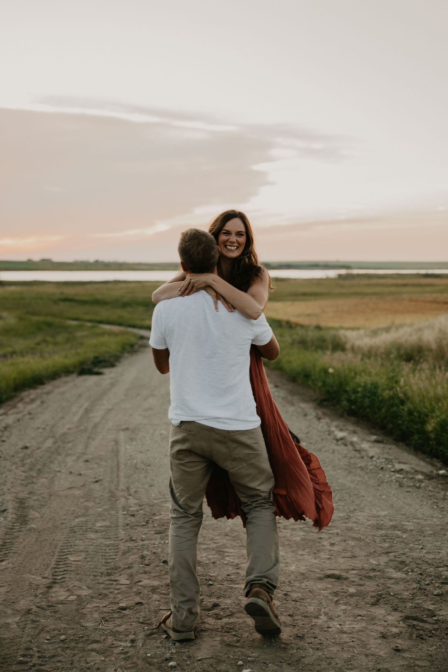 Romantic embrace on rural dirt road during sunset with dramatic sky and grasslands.