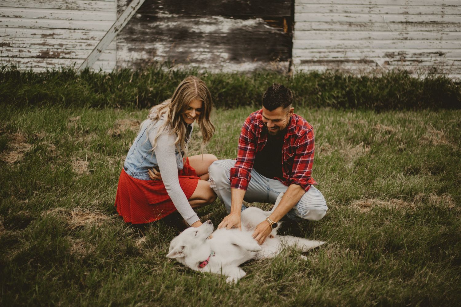 A couple playing with their white dog on the grass near an old barn.