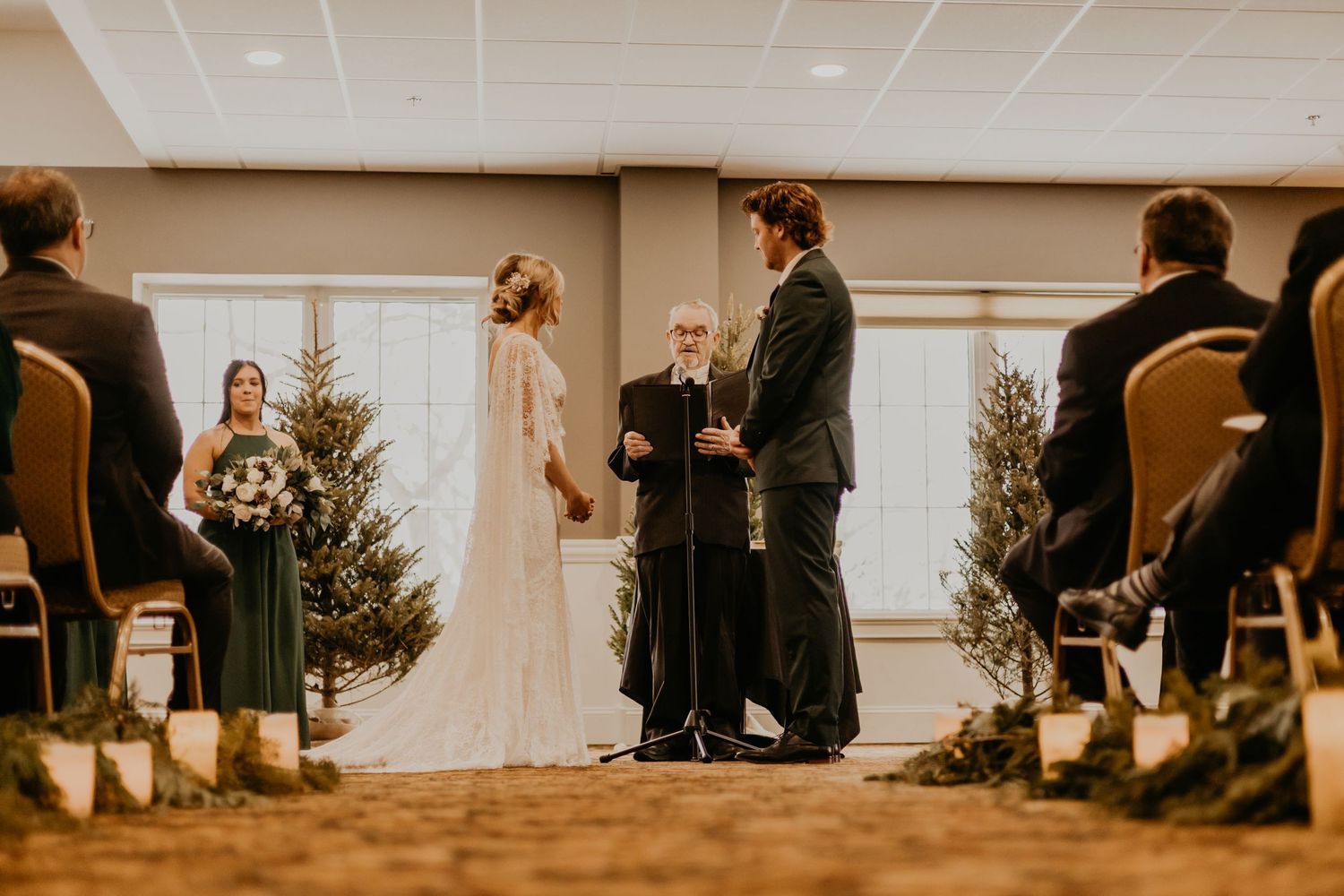 Wedding ceremony in warm-toned room with evergreen trees and guests in attendance.