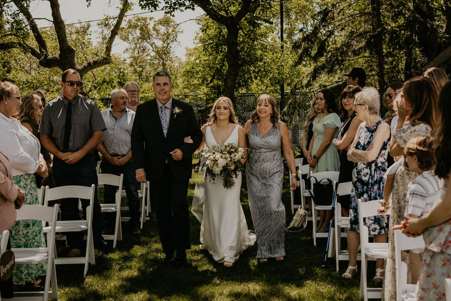 Wedding ceremony guests seated in white chairs on a lush green lawn beneath towering trees.