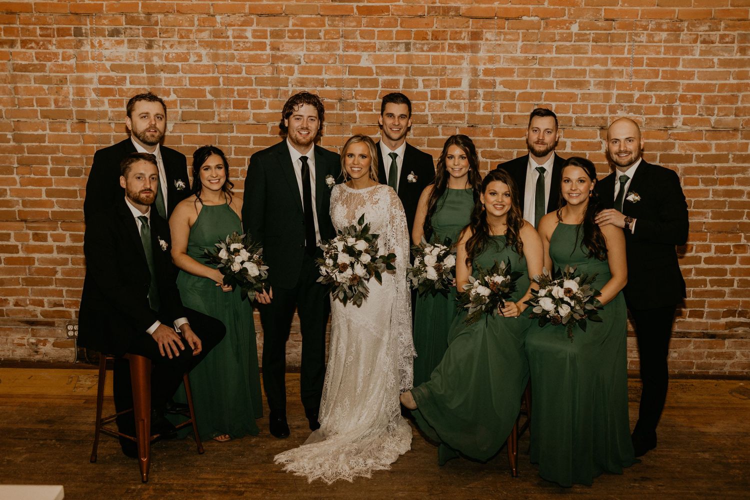 Wedding party poses in front of rustic brick wall with bridesmaids in emerald green dresses and groomsmen in black suits.