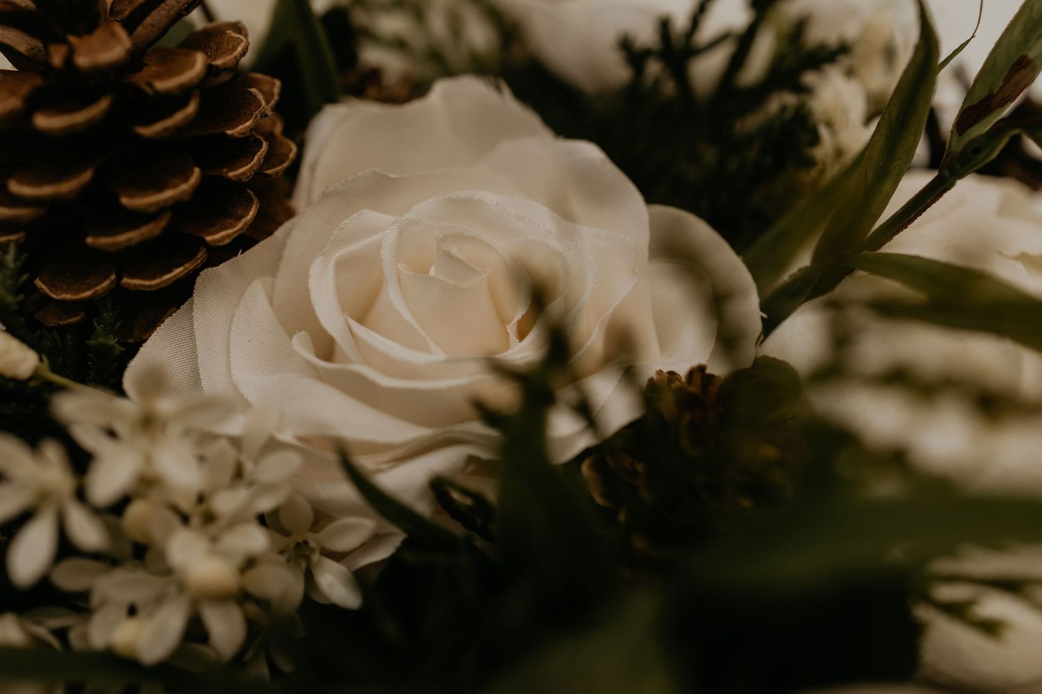 Close-up shot of a white rose and pinecone in a wedding bouquet with moody lighting.