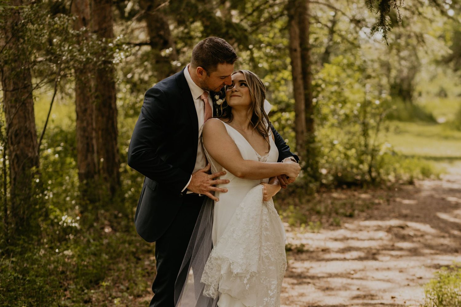 Intimate moment between couple on woodland path in elegant wedding attire.
