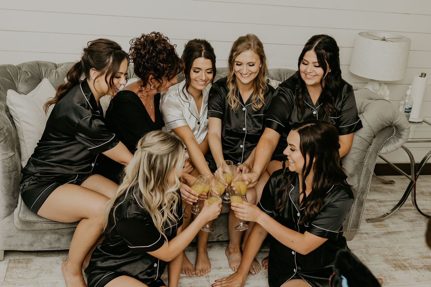 Group wearing matching silk robes toast together on a gray couch during pre-wedding celebrations.
