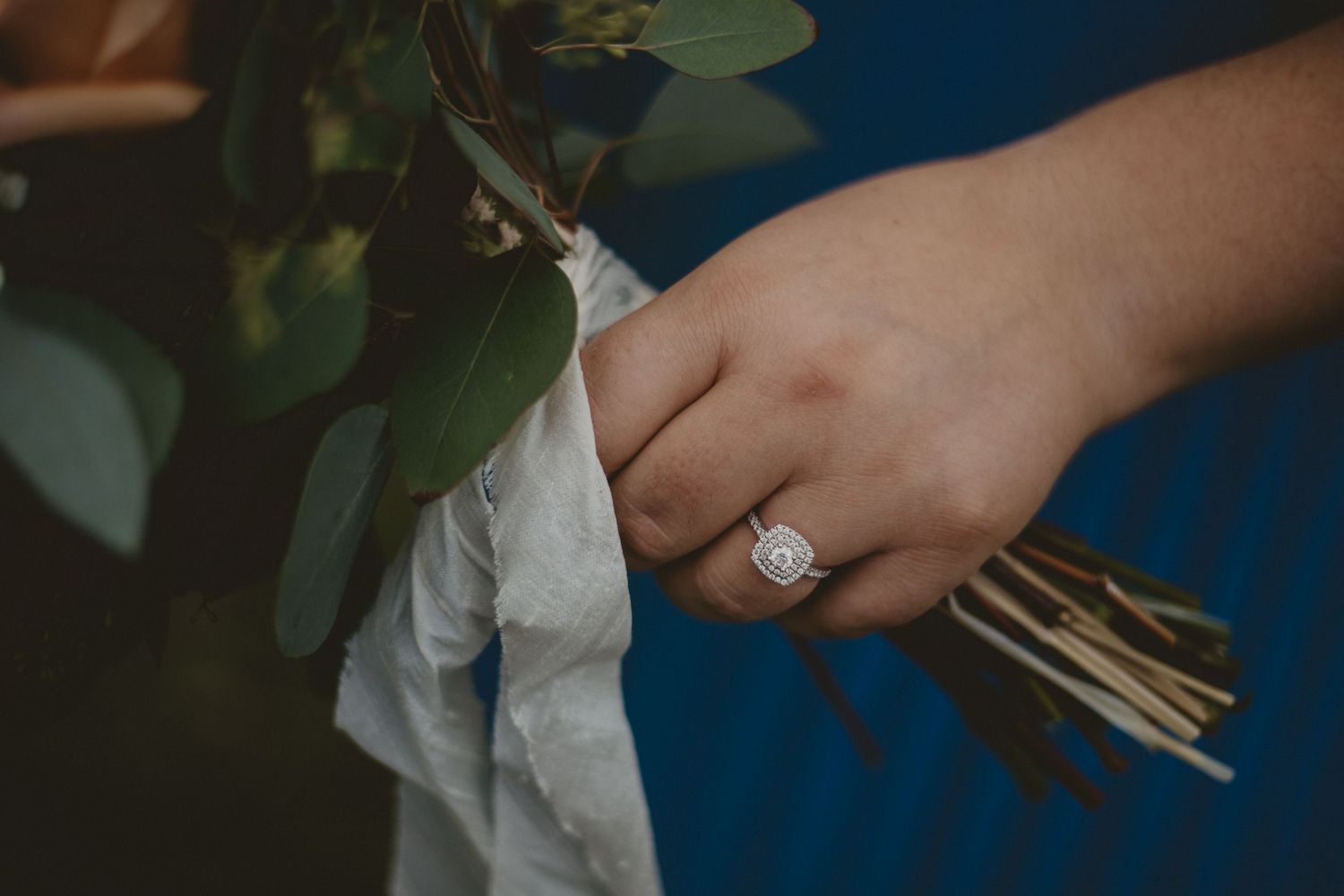 Close-up of diamond engagement ring while holding wedding bouquet with greenery.