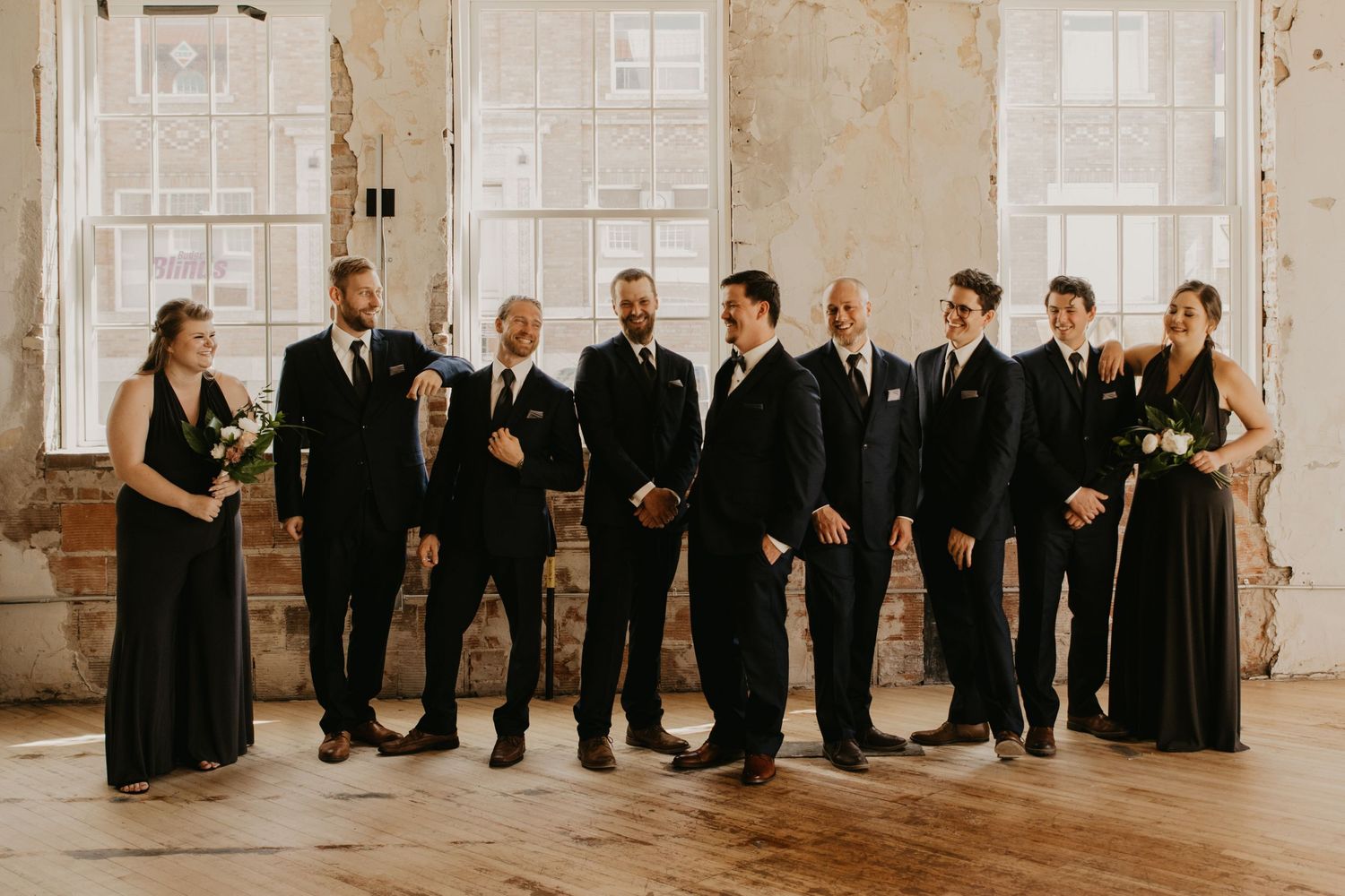 Wedding party in black formal attire poses together in an elegant industrial space with large windows.