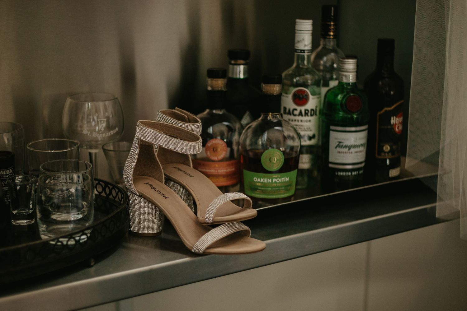 Wedding shoes rest on a bar shelf next to various spirits and glassware.