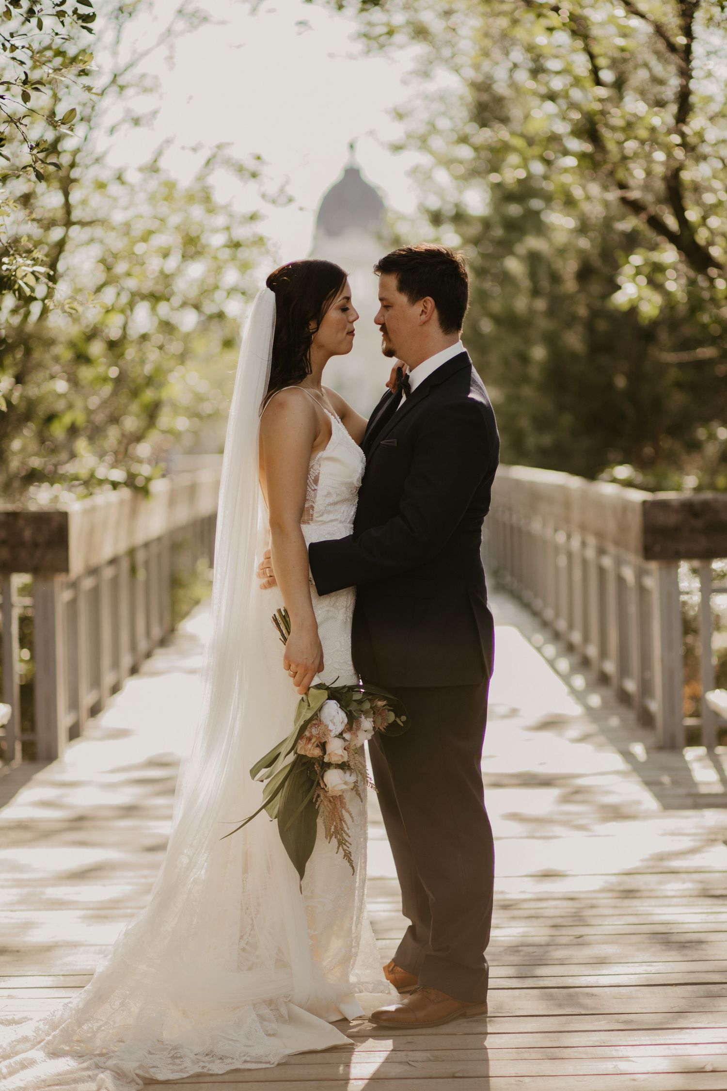 Wedding couple in romantic pose on a wooden bridge with cathedral dome visible in background.