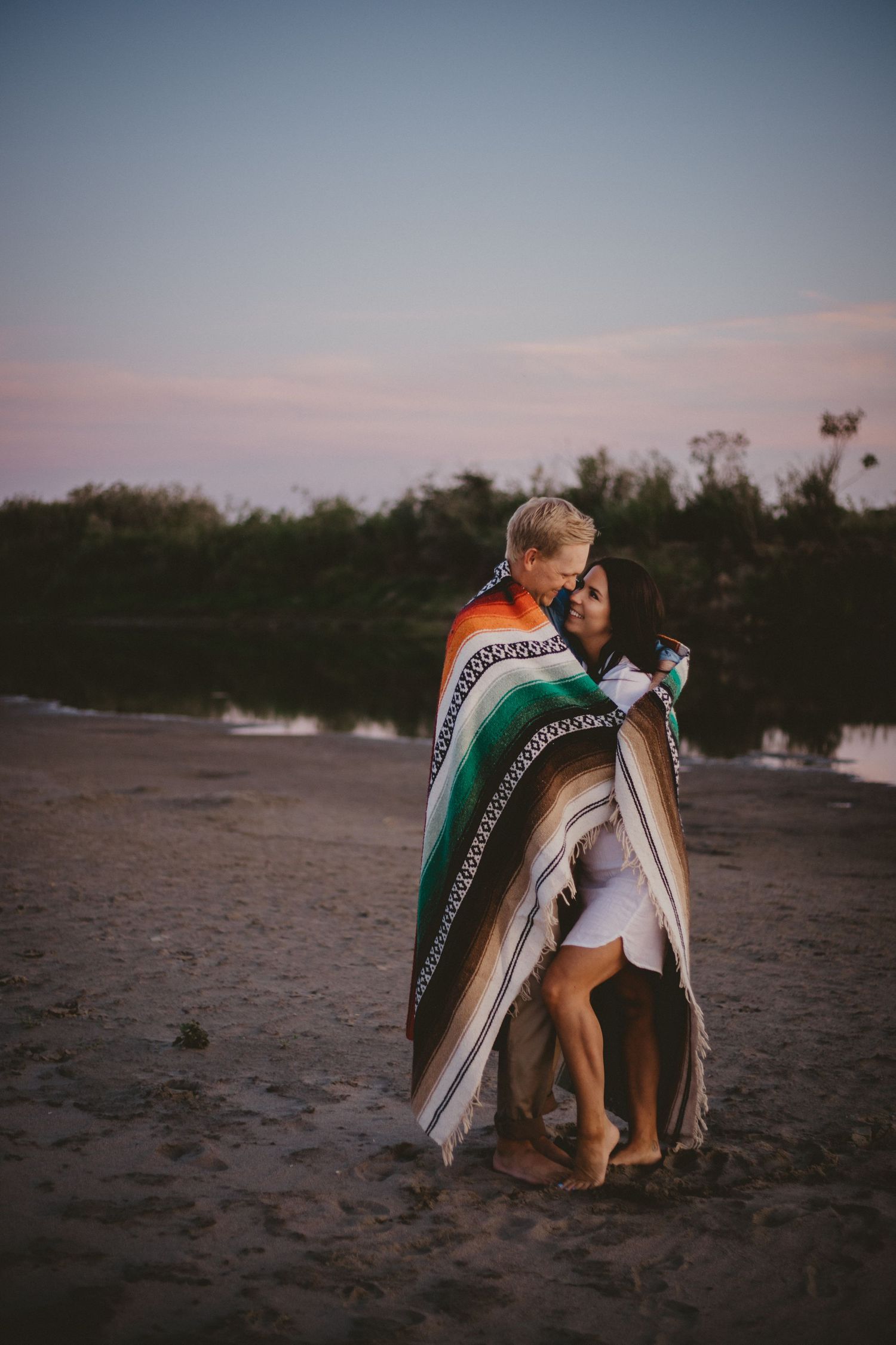 A couple wrapped in a striped blanket embraces on a beach at sunset.