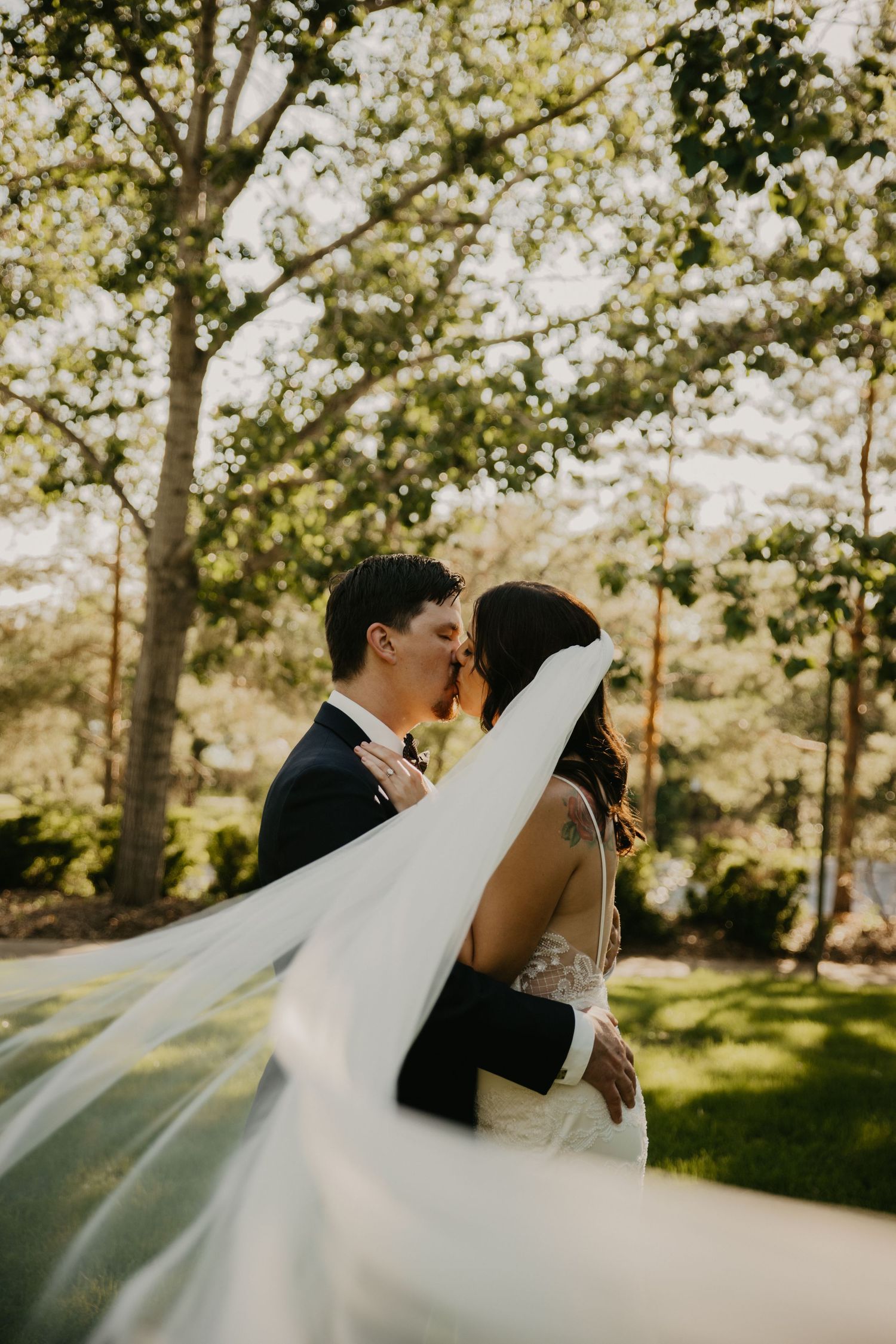 Romantic wedding portrait with flowing veil in sunlit garden setting.