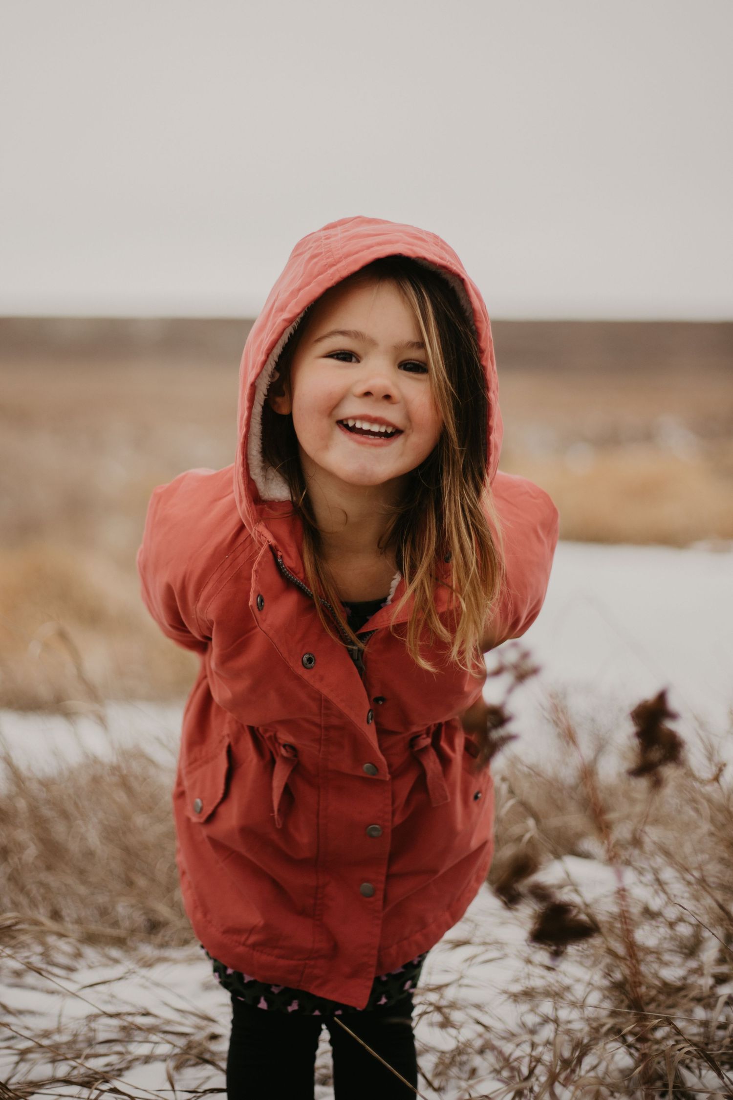 A child in a coral-colored hooded jacket plays outdoors in a snowy winter field.