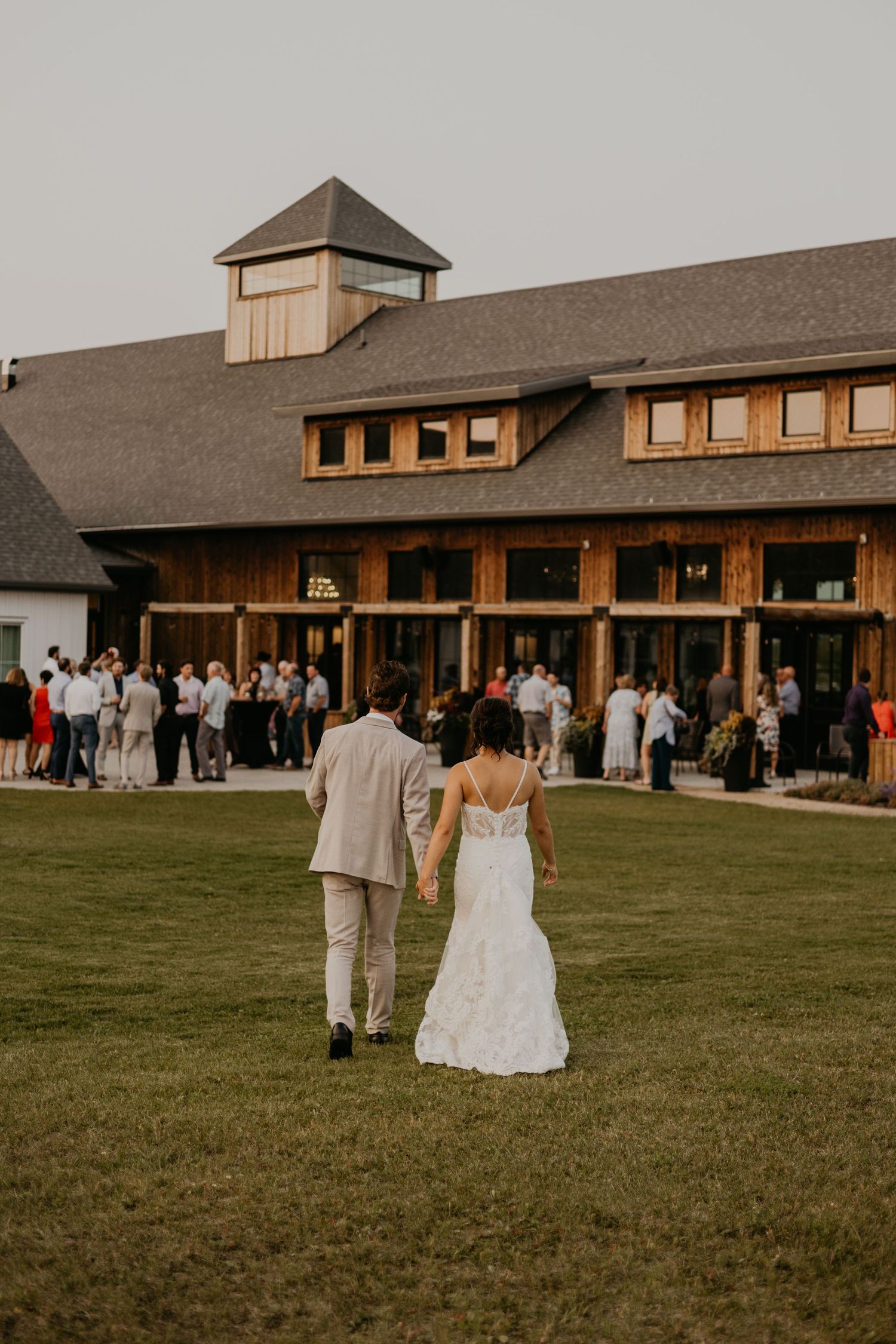 A couple walks on the lawn toward a rustic wedding venue as guests gather outside the building.