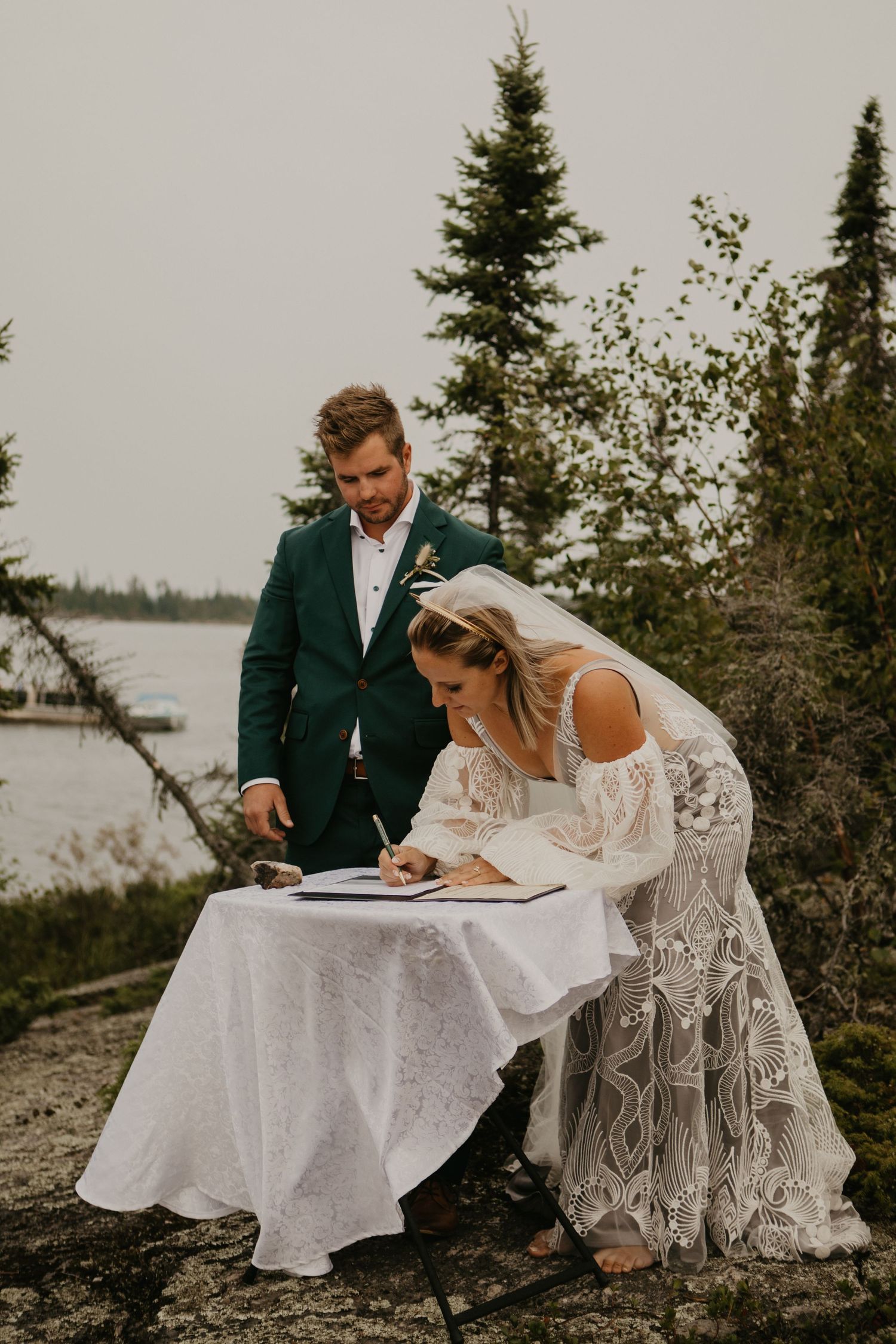 A couple signing wedding documents on a white-clothed table by a scenic lake.