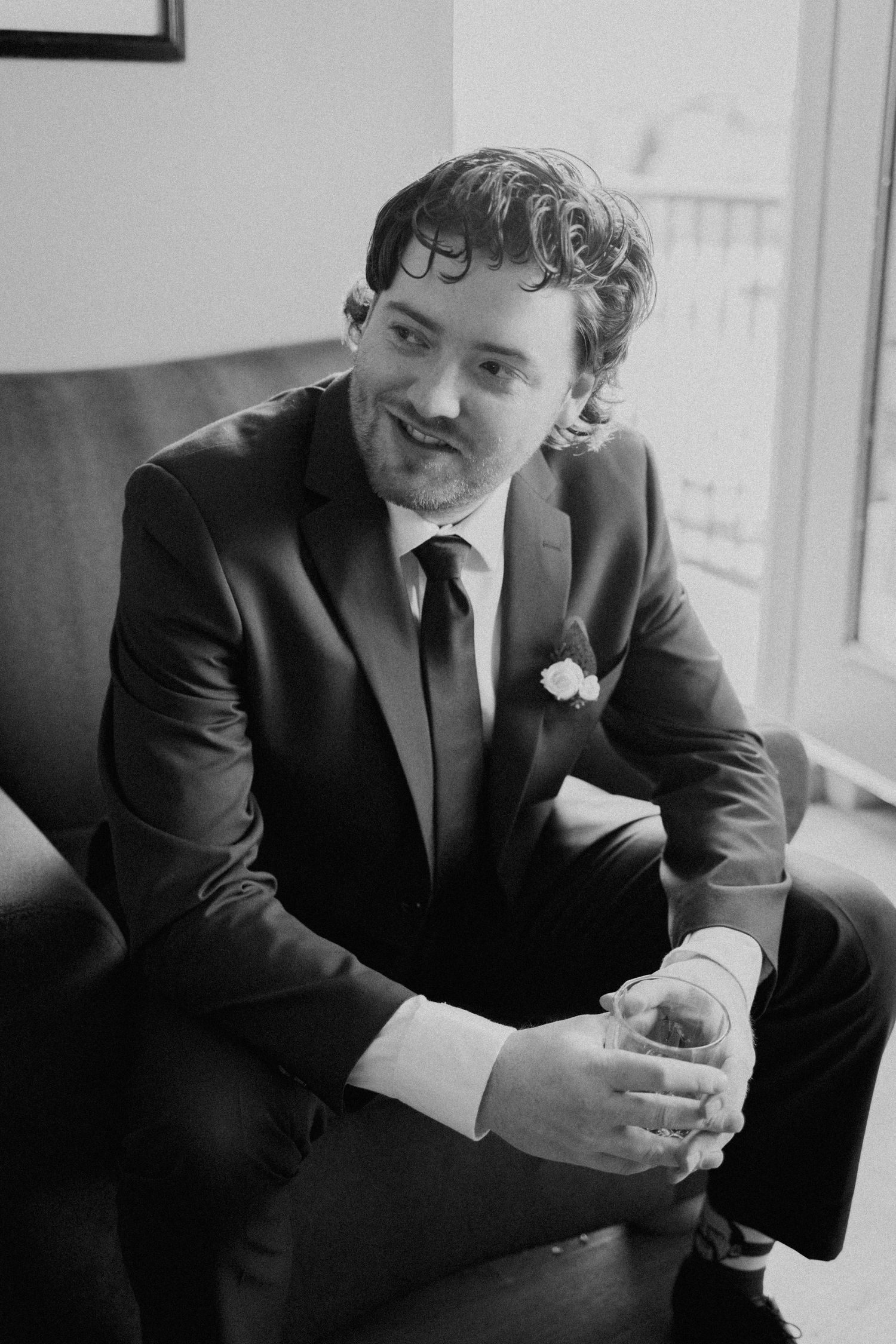 A stylishly dressed gentleman in a dark suit holding a glass while sitting near a window in black and white.
