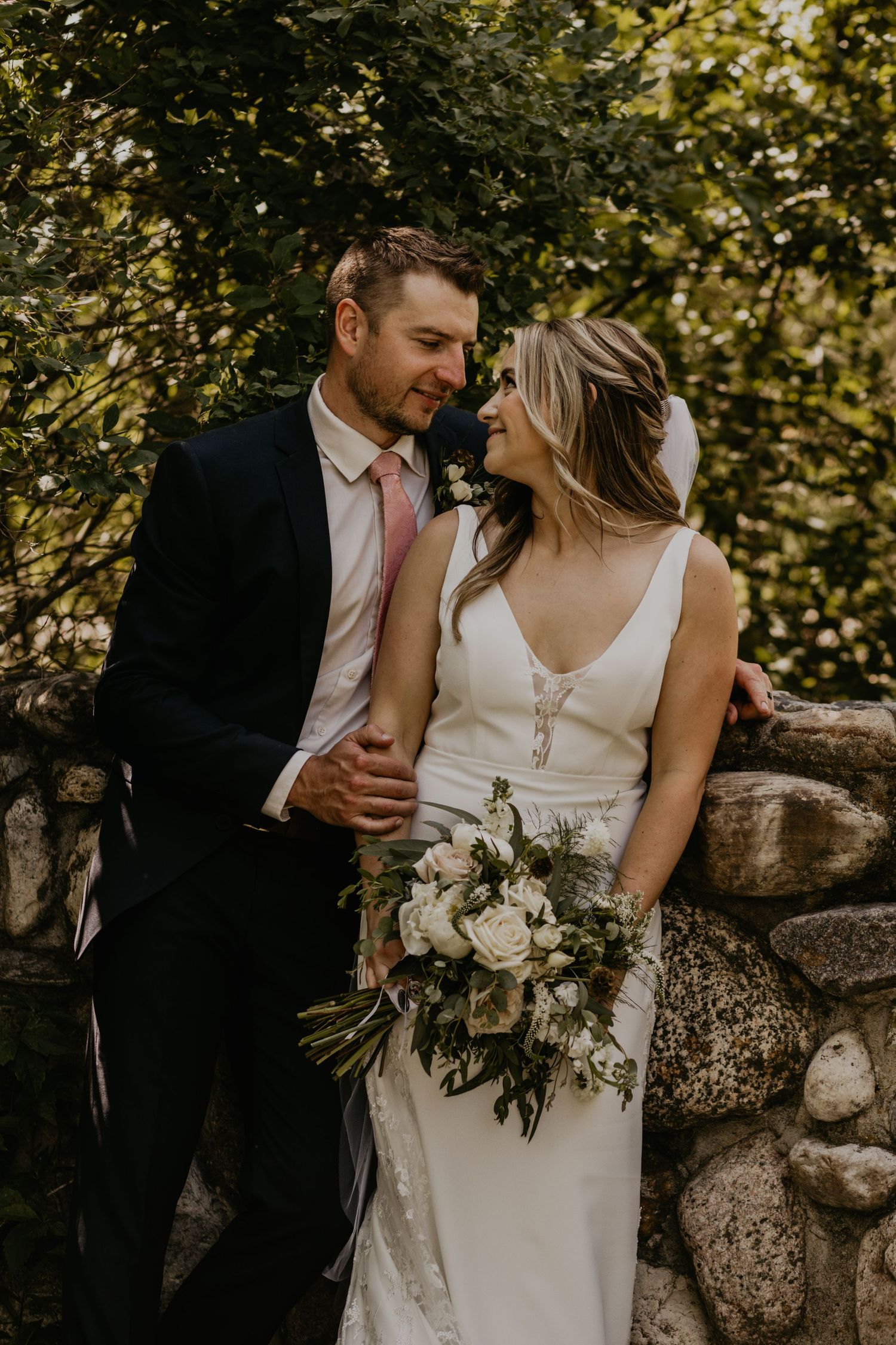 Wedding couple sits on stone wall sharing intimate moment with bride holding elegant bouquet.