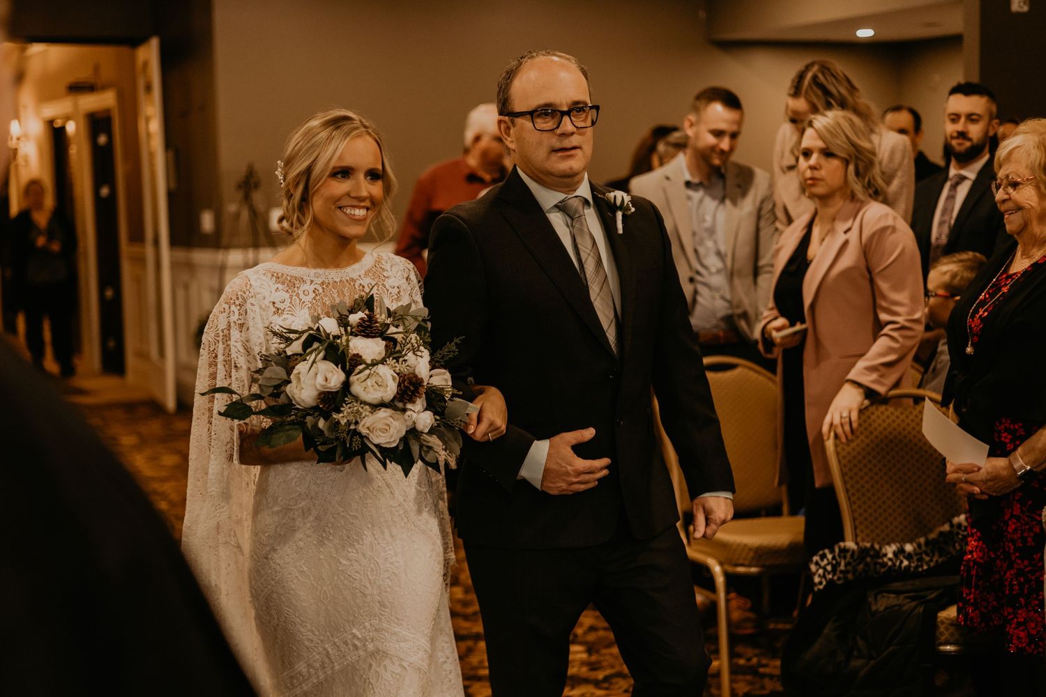 A bride in white lace holds a bouquet while walking down the carpeted aisle during the ceremony.
