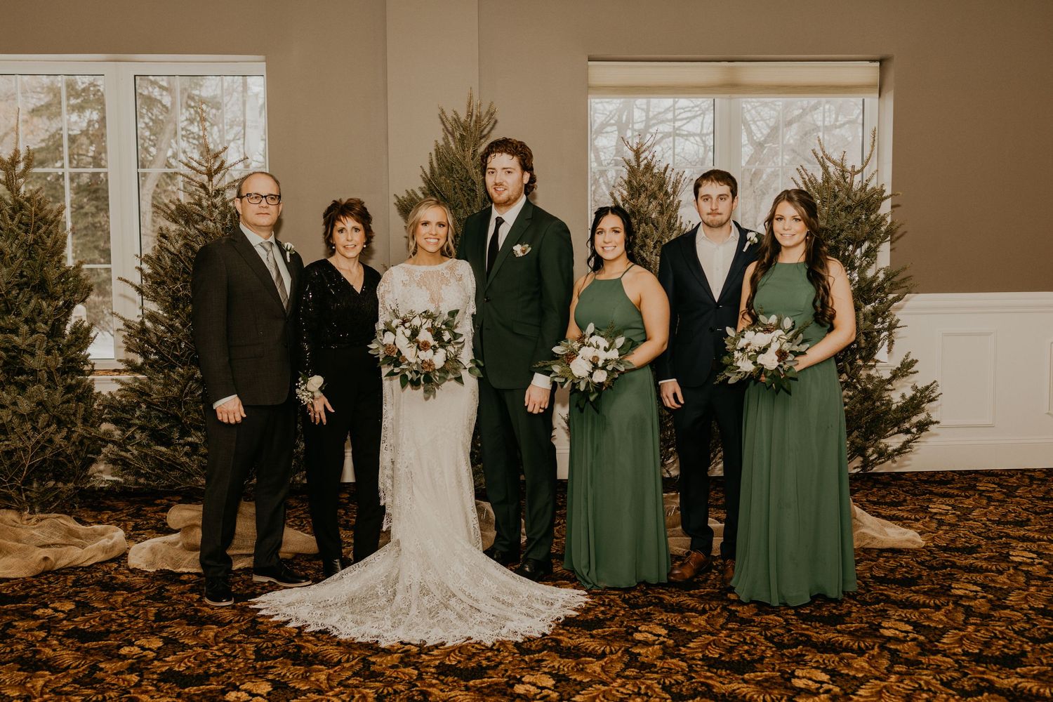 Wedding party in black and emerald green attire poses for formal photos with decorative evergreens.