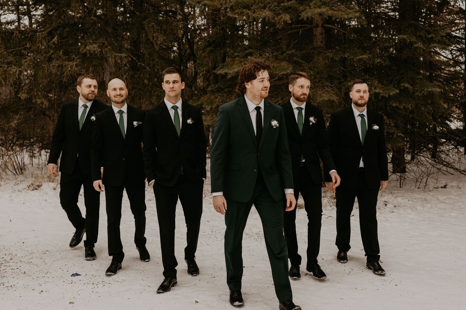 Groomsmen walking in formation through snow wearing matching black suits.
