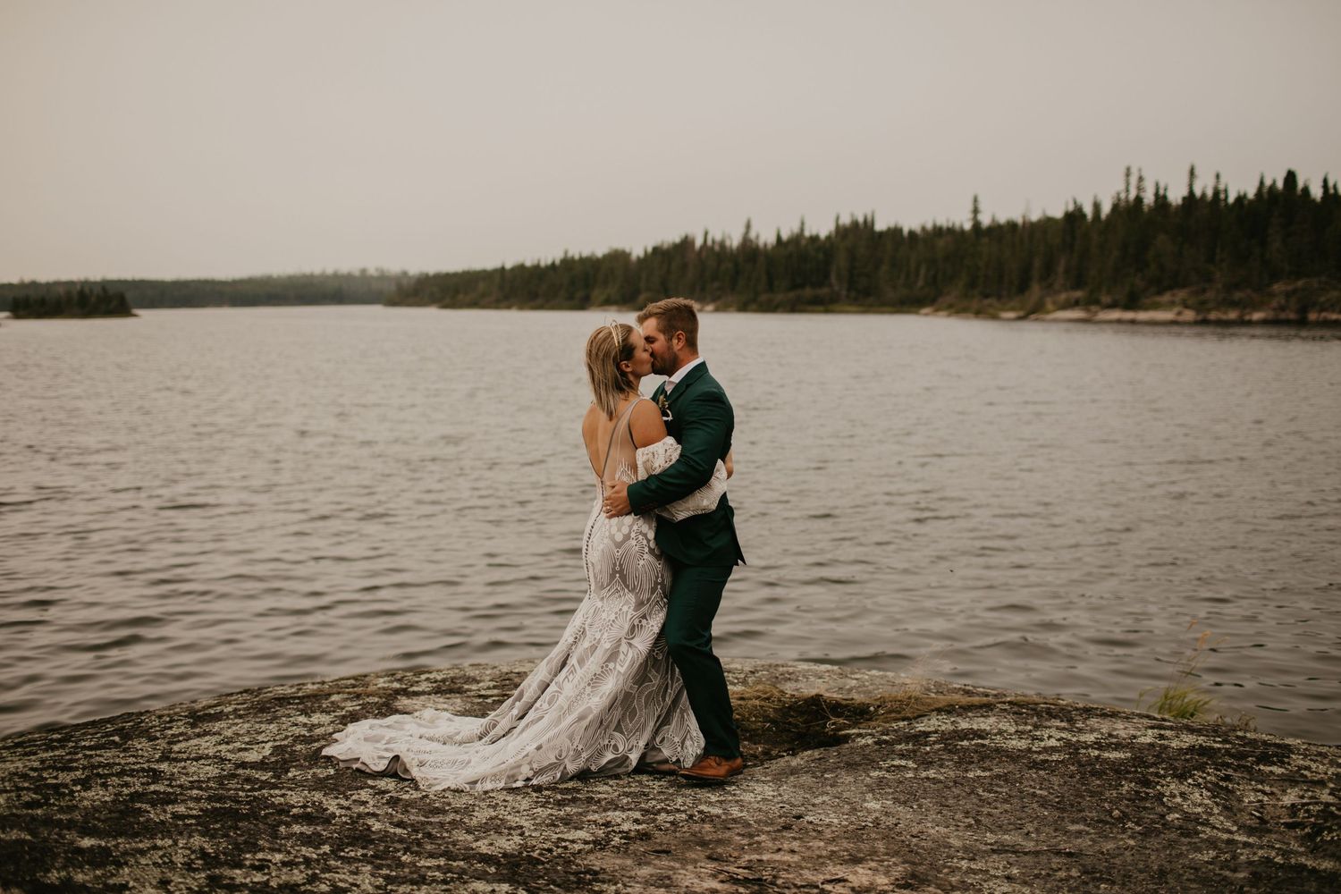 A tender moment between newlyweds on a rocky shore with a serene lake in the background.