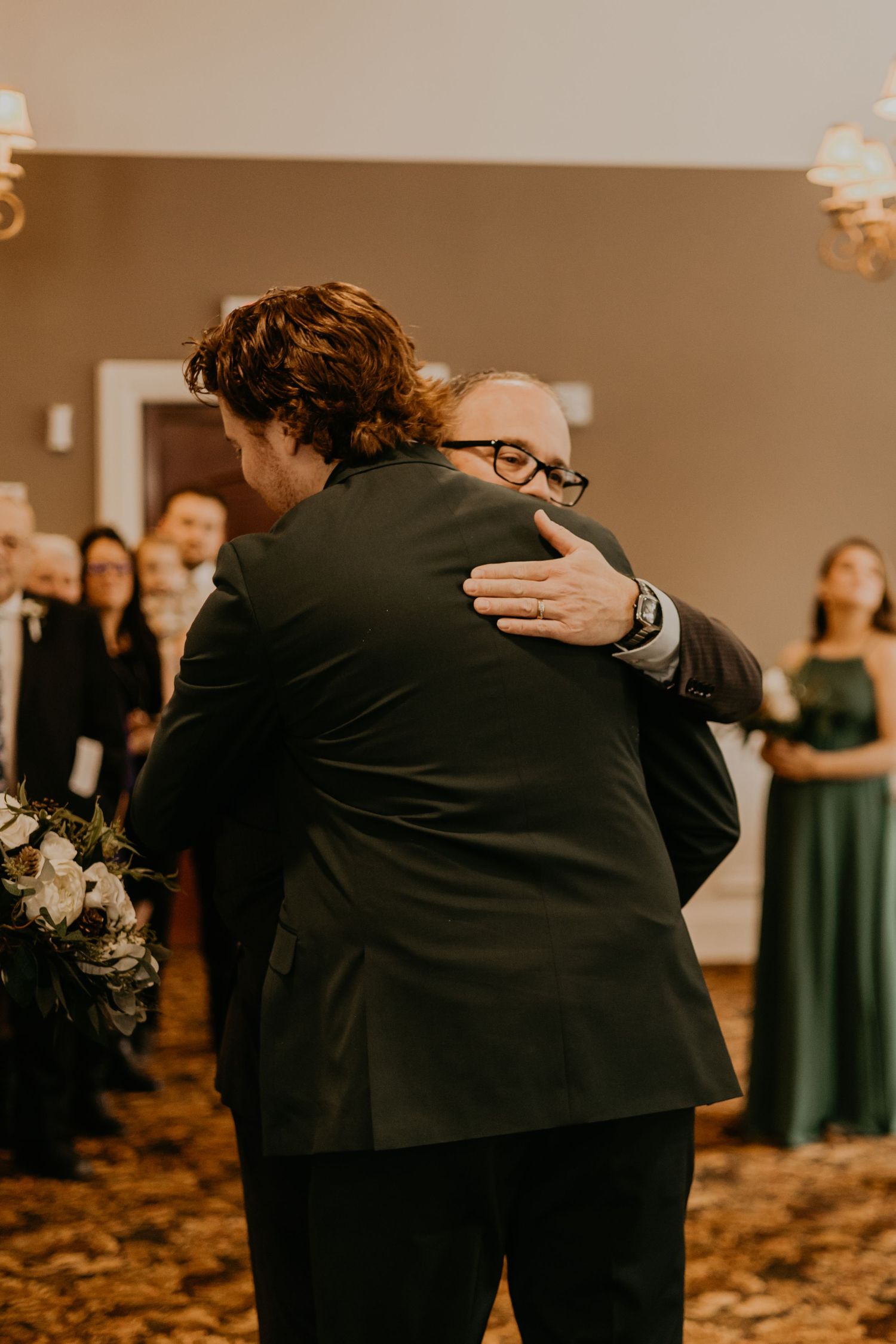 Two people share a warm embrace during a wedding ceremony as guests look on in the background.