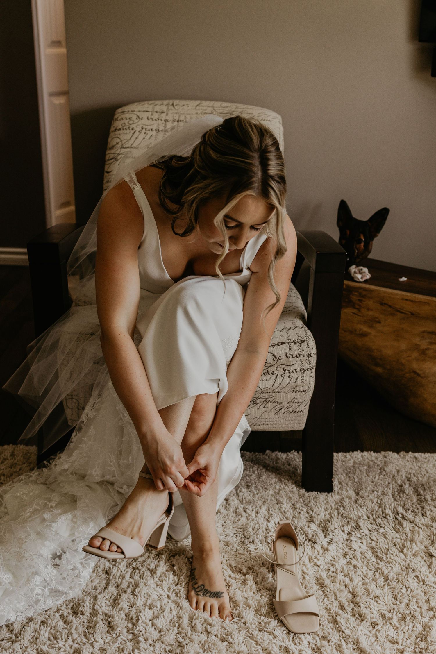 Bride in white dress puts on wedding shoes while sitting in chair with black cat in background.