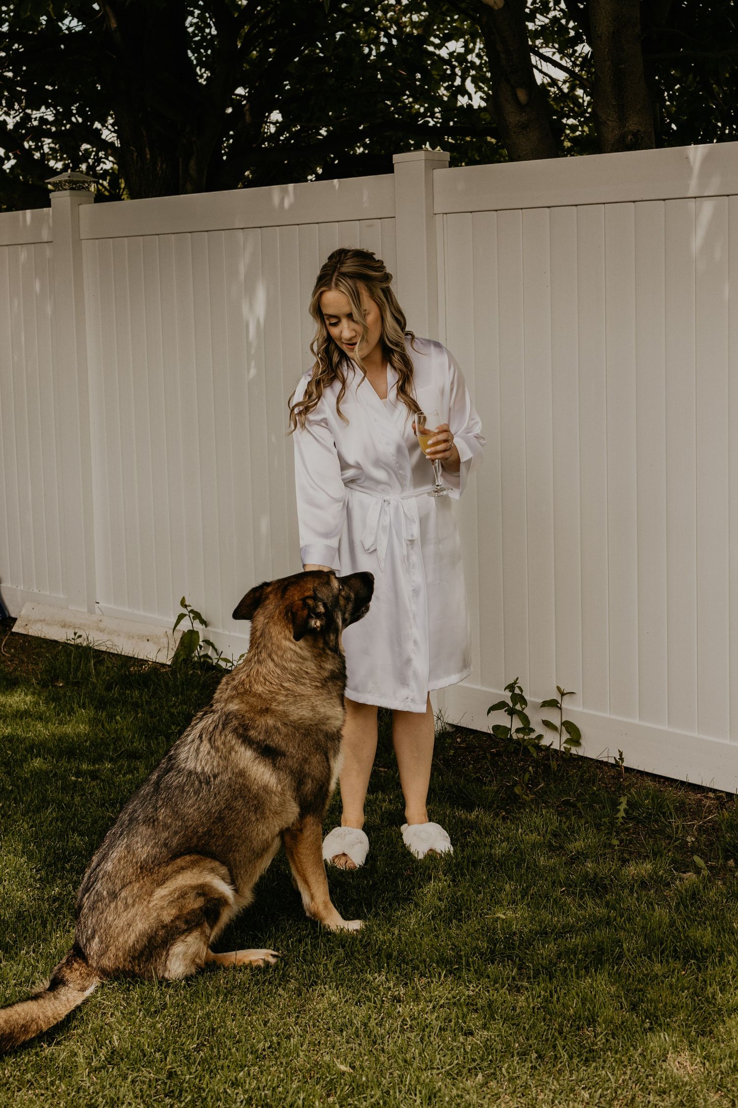 Bride in white robe stands with German Shepherd dog by white fence during wedding preparations.