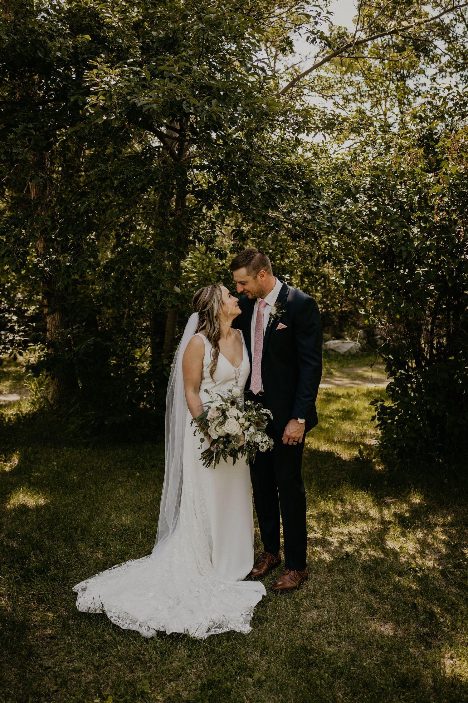Full-length wedding portrait with bride in flowing train dress against natural garden backdrop.