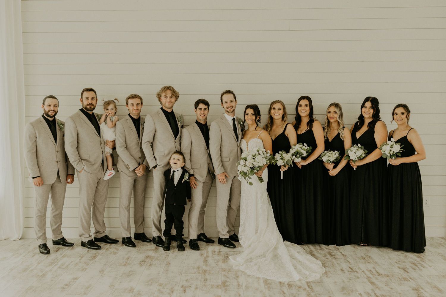 Large wedding party with bridesmaids in black and groomsmen in tan suits pose against white backdrop.