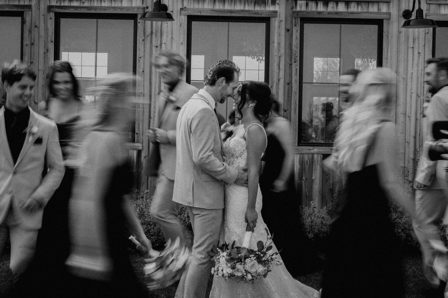 Wedding celebration with motion-blurred guests dancing around the couple outside a wooden venue.