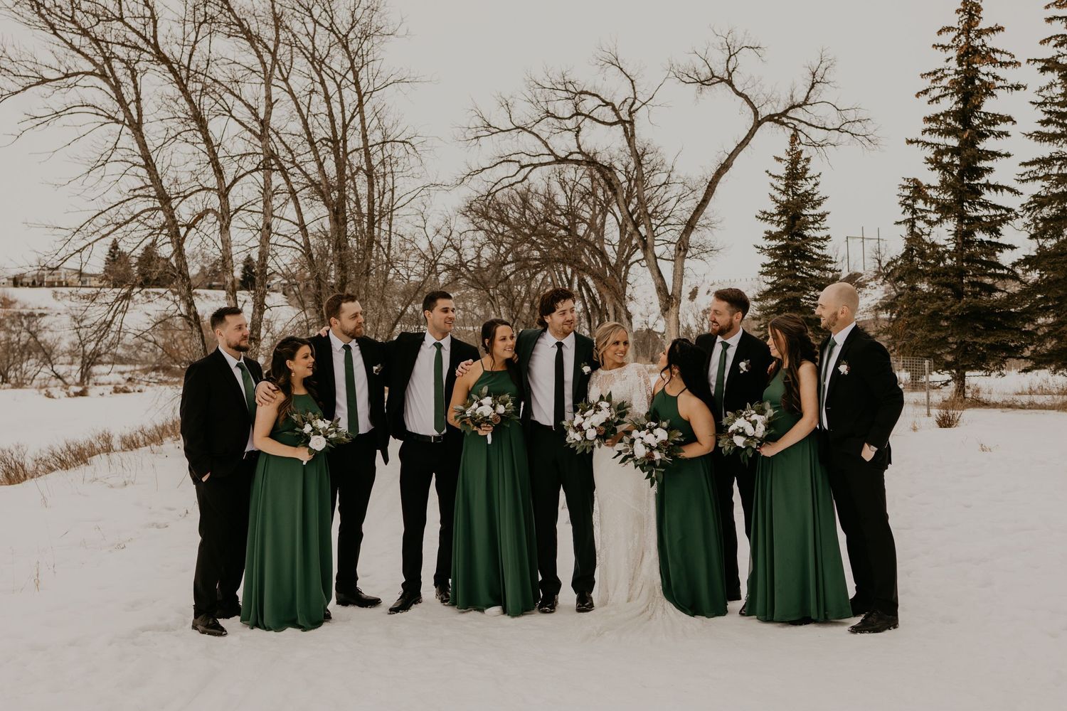 Wedding party in formal attire poses for photos in a winter landscape with bare trees and snow.