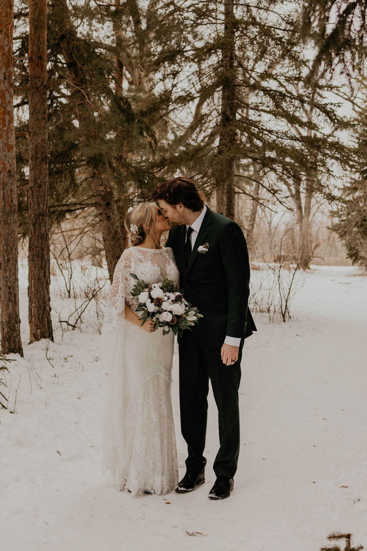 A winter wedding portrait of a couple holding a bouquet among snow-covered pine trees.